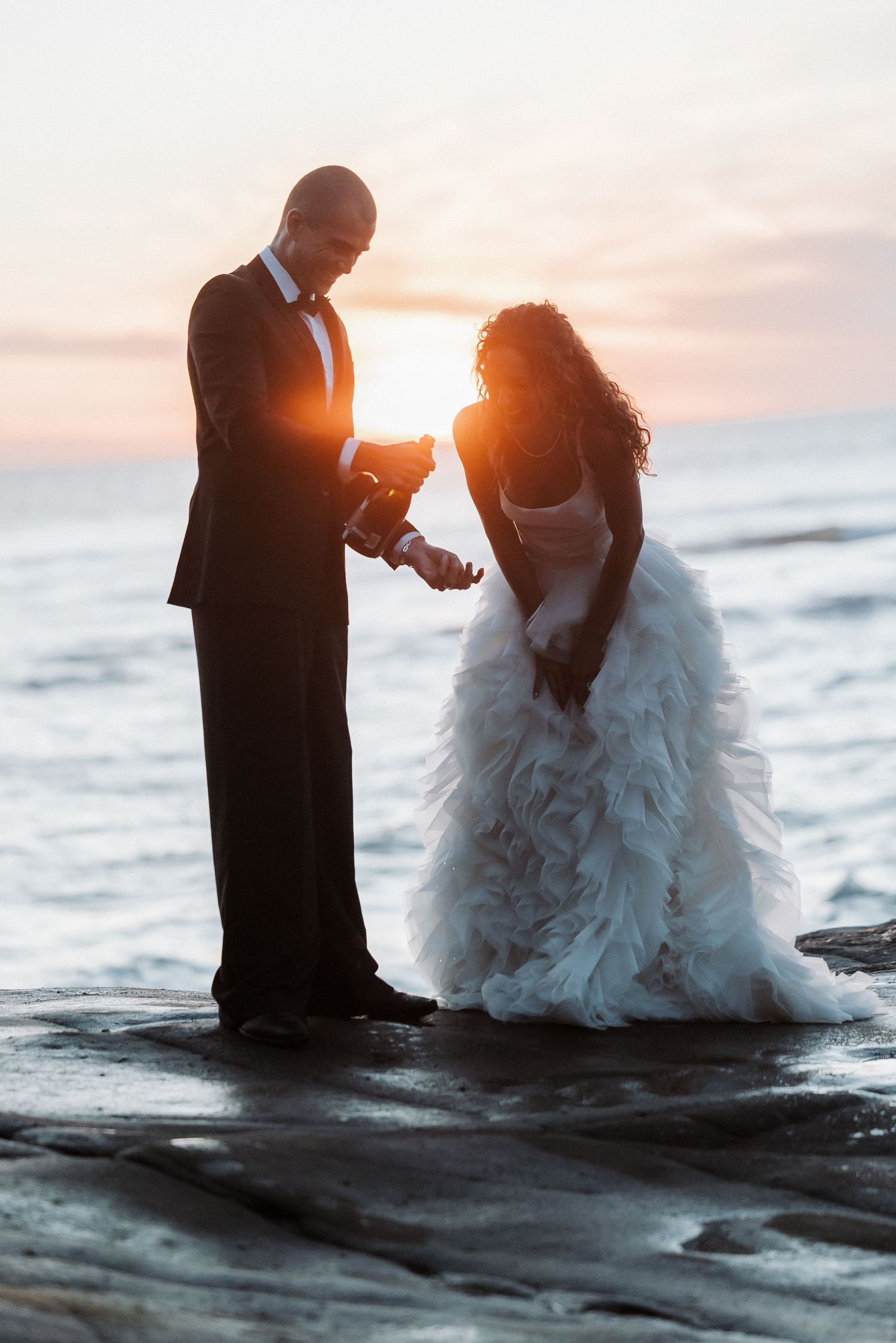 Wedding couple shares an intimate moment at sunset on rocky beach as waves crash behind them.