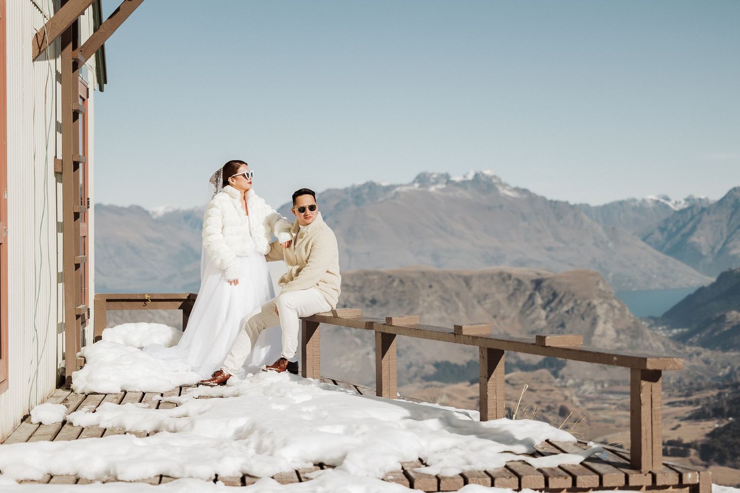 Couple in wedding attire standing on wooden deck overlooking majestic mountain vista.