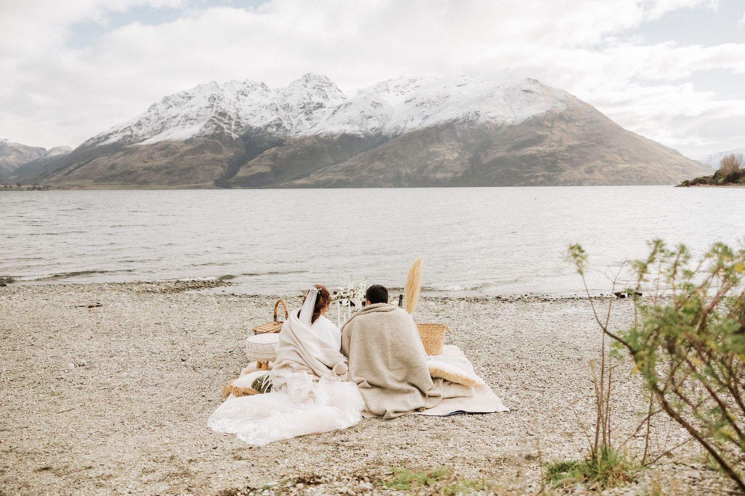 A couple in white sits on a picturesque mountain lakeside with dramatic snow-capped peaks in the background.