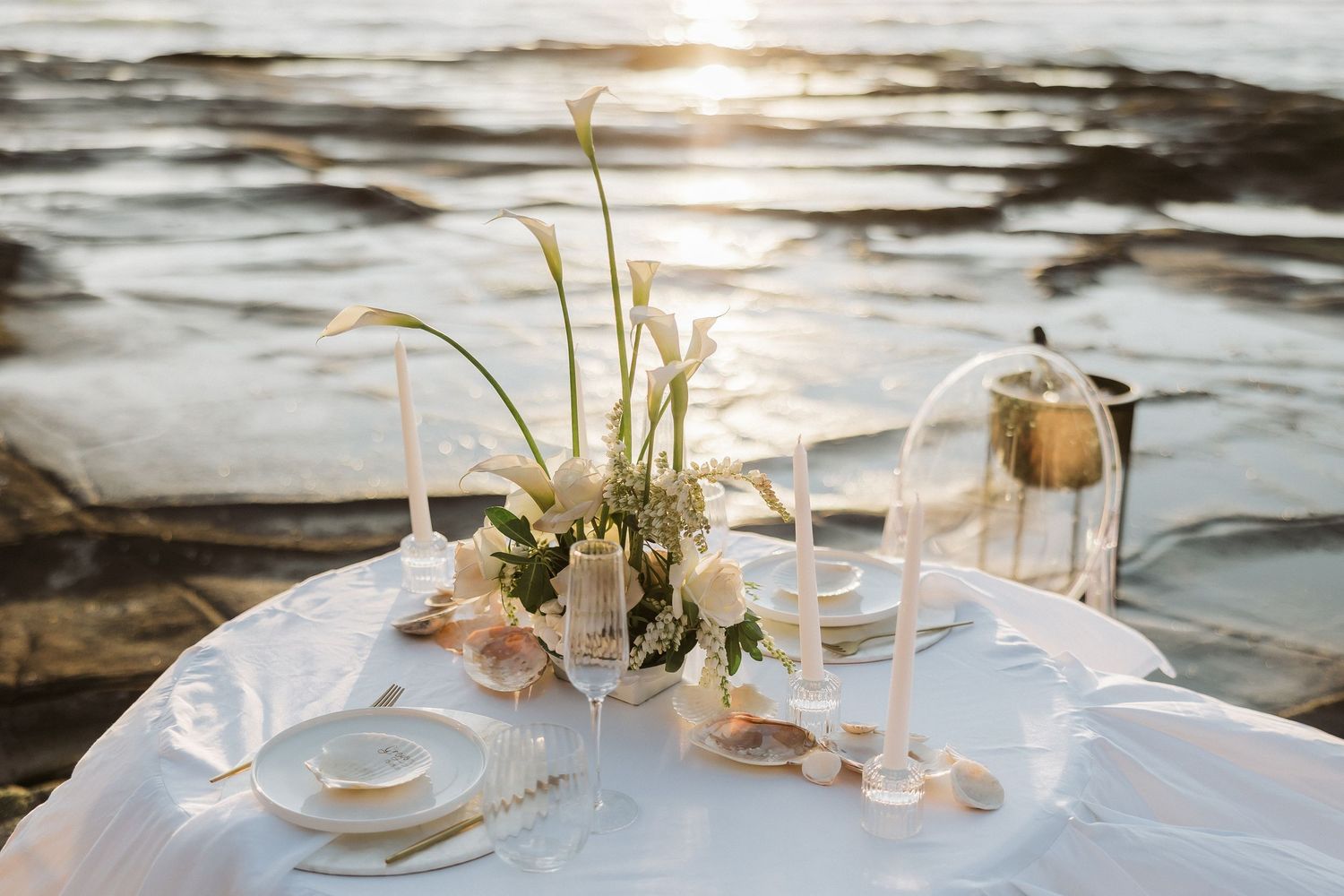 Elegant beach wedding reception setup with white tablecloths and floral centerpieces at sunset along the ocean shore.