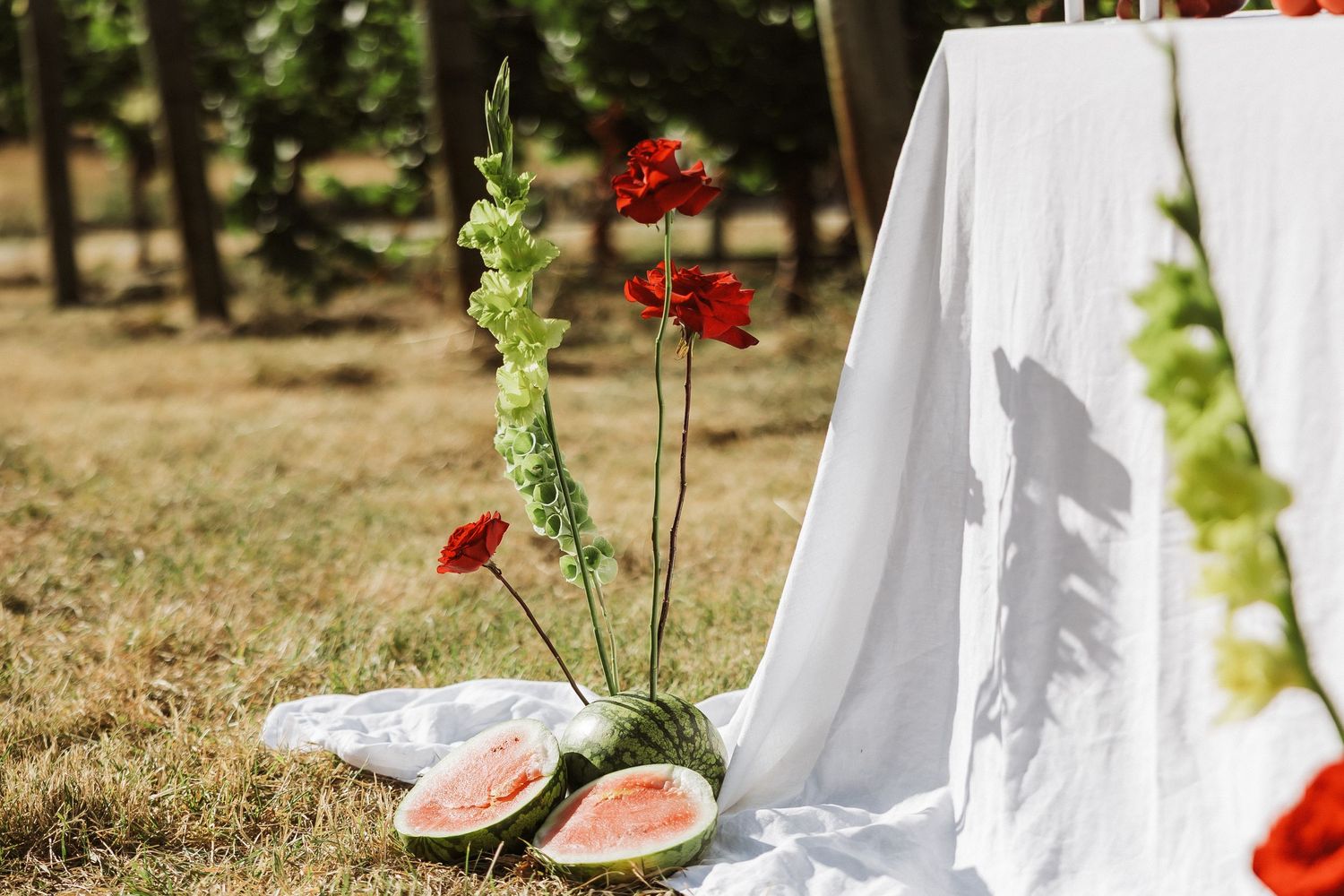 Elegant outdoor table setting with red roses and watermelon slices on white linens in a sunny garden setting.