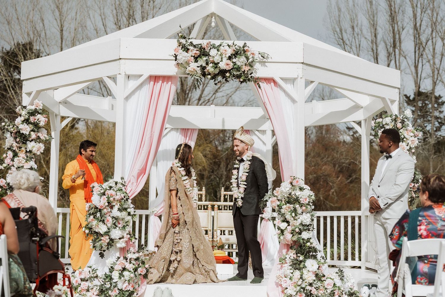 A white wedding gazebo decorated with pink and white drapes and floral arrangements in an outdoor setting.