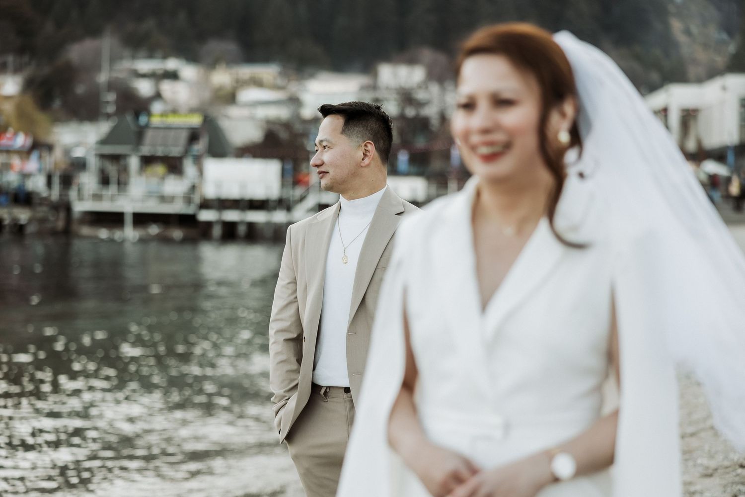 A bride and groom share a joyful moment by the waterfront with boats and houses in the background.