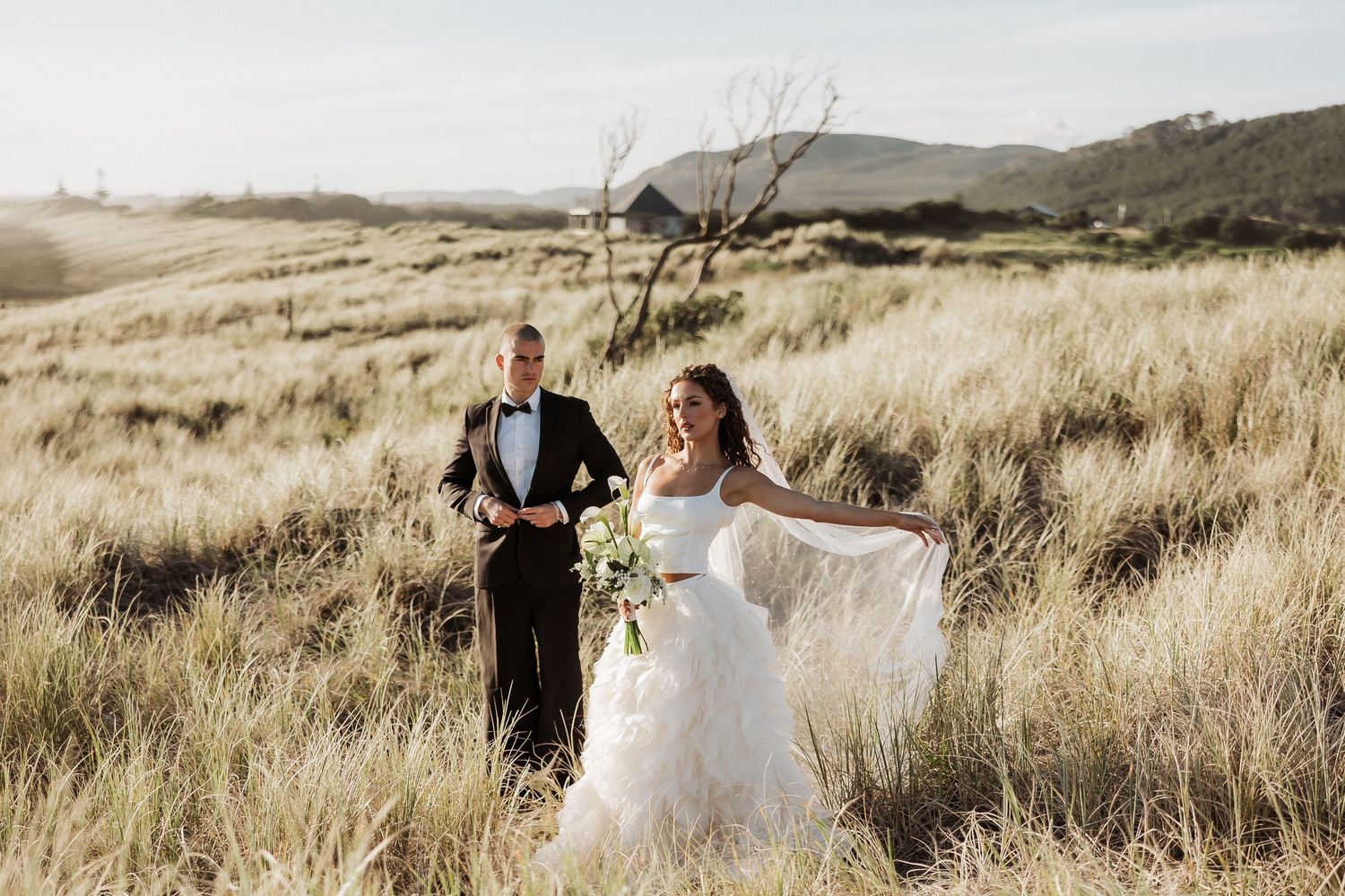 Wedding couple poses in a scenic field with mountains and dried grass in the background.