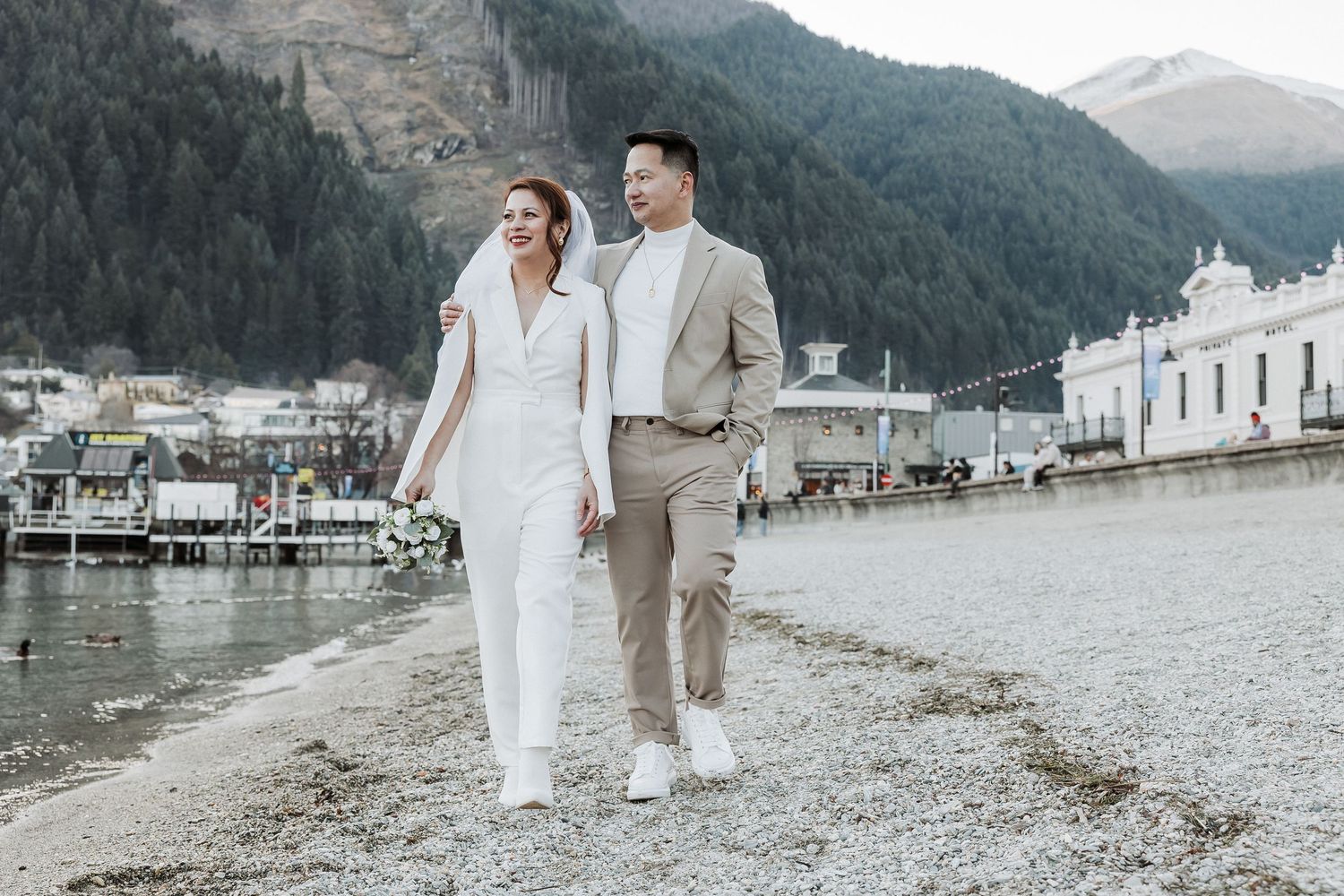 Couple strolling along scenic lakeside beach with mountain backdrop in muted tones.