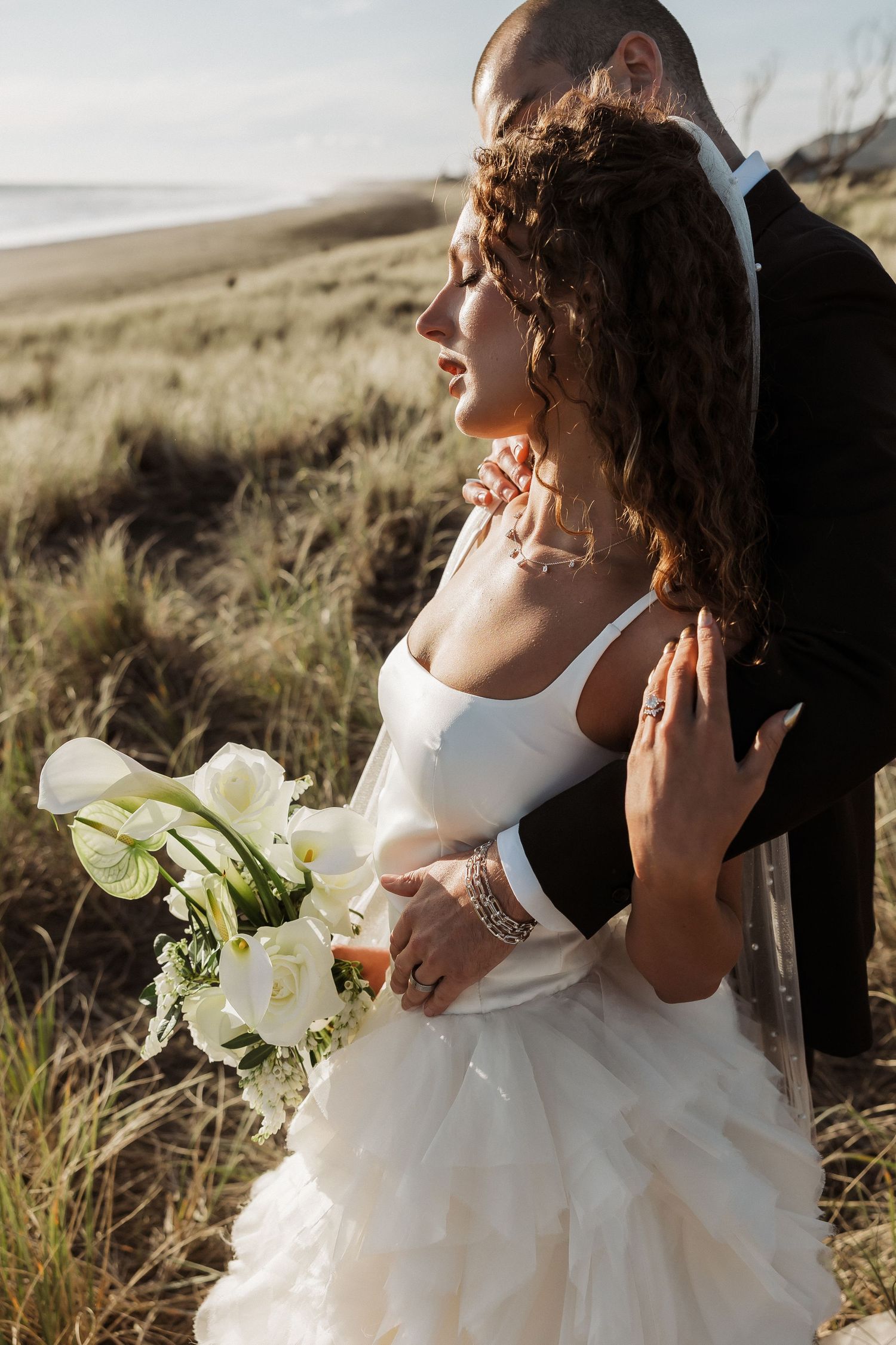 A bride in a white wedding dress holds a bouquet of white calla lilies against a scenic outdoor backdrop.