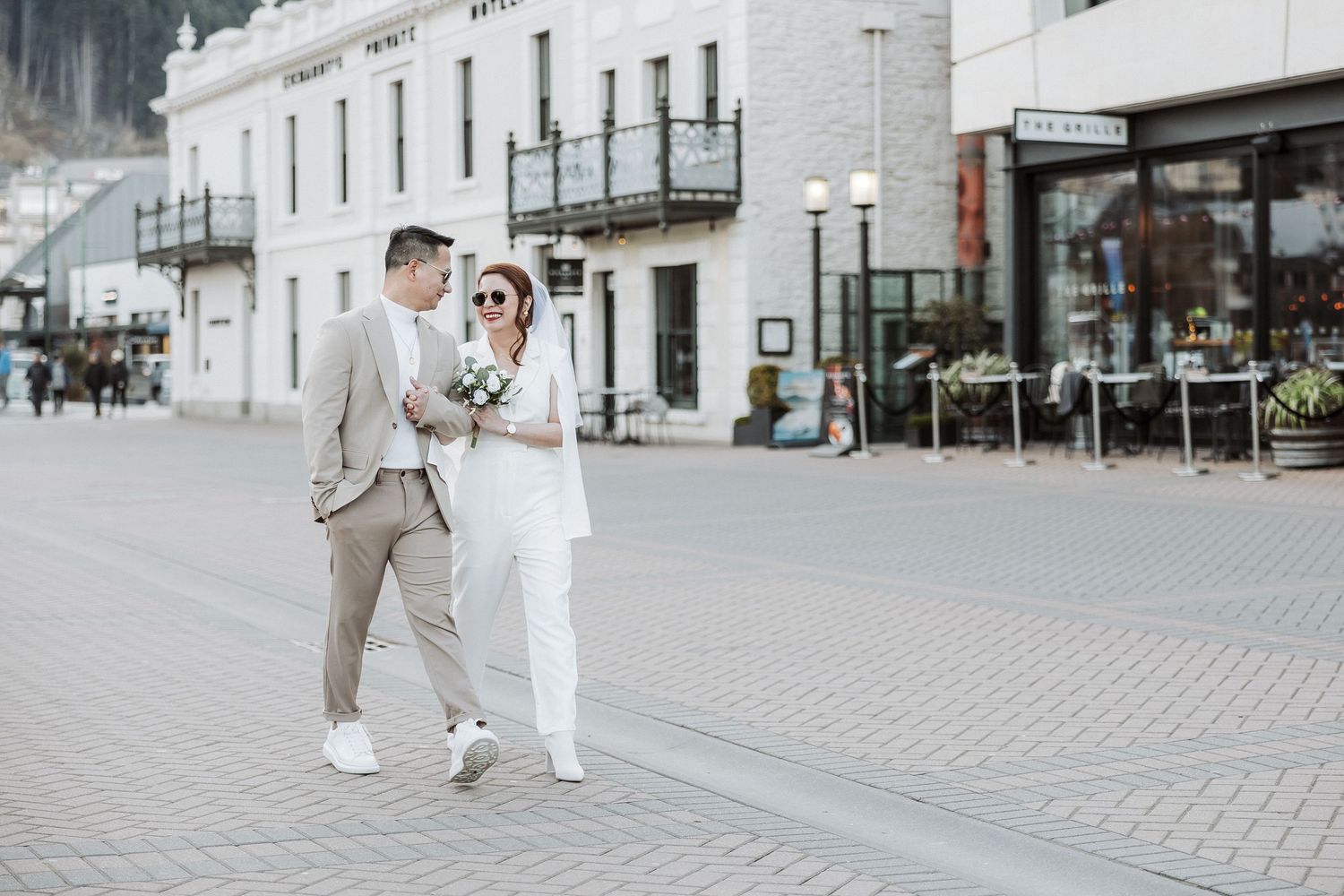 A couple in elegant wedding attire walks together on a cobblestone street in an urban setting.