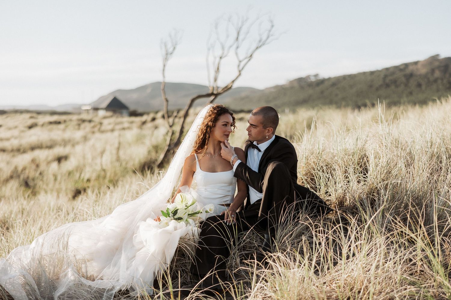 Wedding couple embracing in a field of tall grass with mountains and dramatic sky in the background.