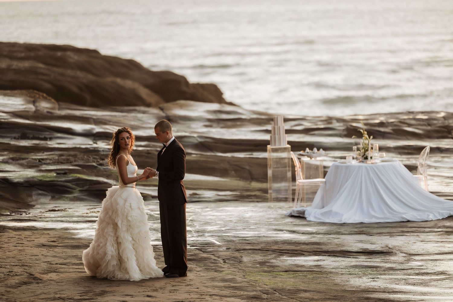 Newlyweds share romantic moments along a rocky coastal beach at sunset with waves crashing in the background.
