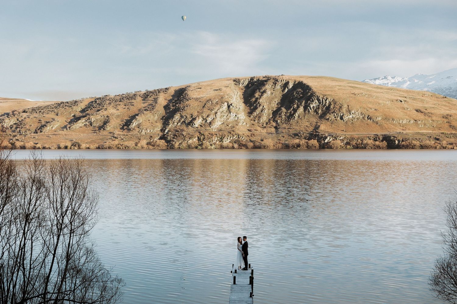 A panoramic view of a tranquil lake surrounded by golden hills and mountains under a bright blue sky.