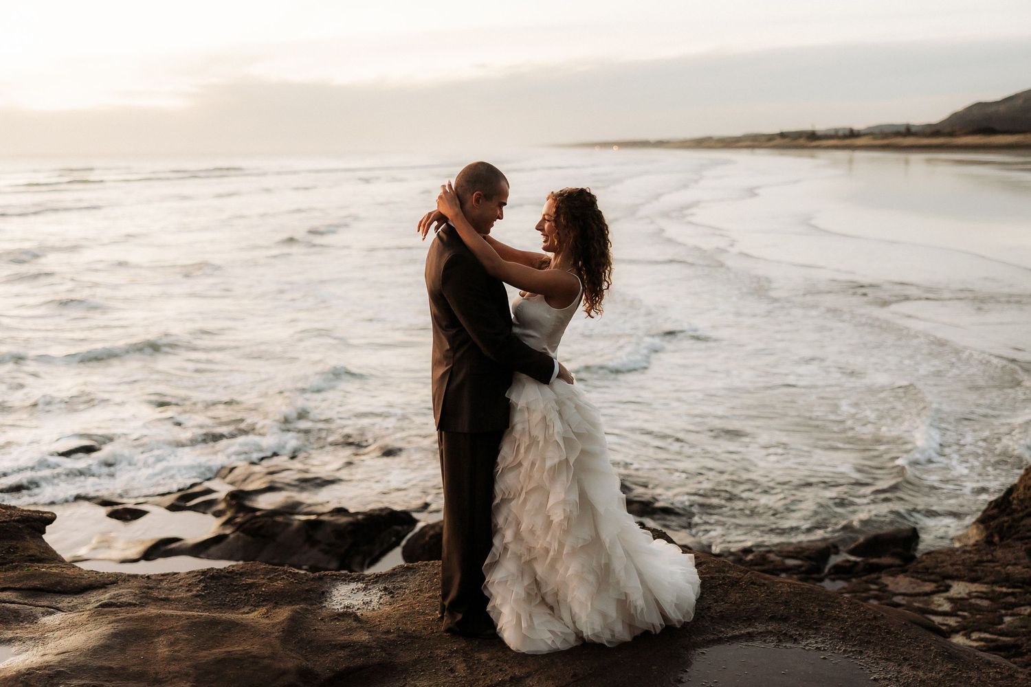 Romantic couple embraces on a rocky beach at sunset with waves crashing in the background.