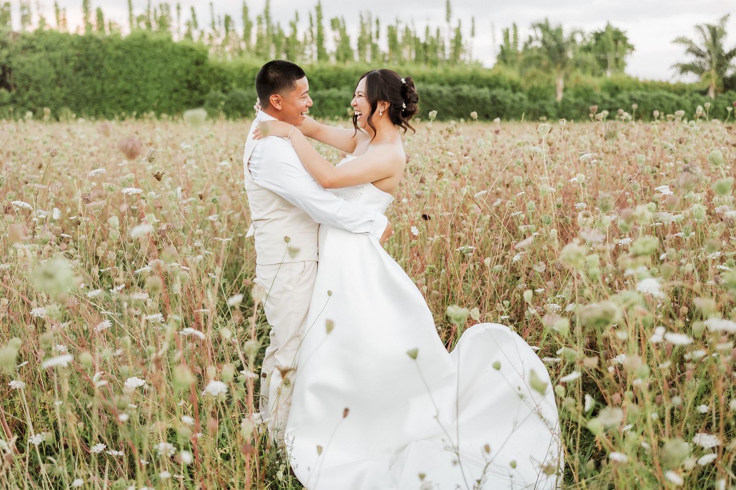 A couple in white outfits share romantic moments among wildflowers in a scenic outdoor photoshoot.