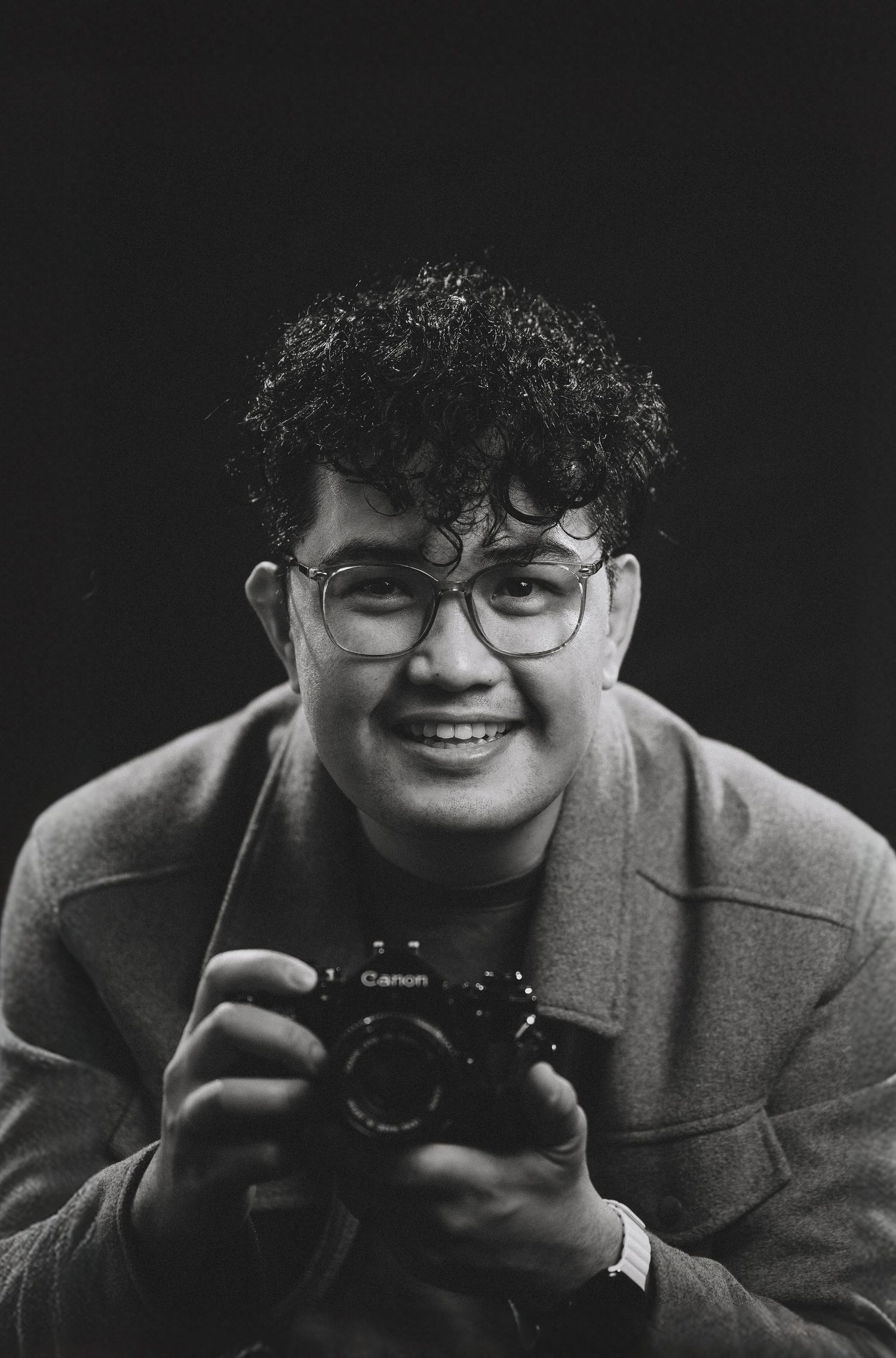 Portrait of photographer in grey blazer holding camera and smiling against black background.