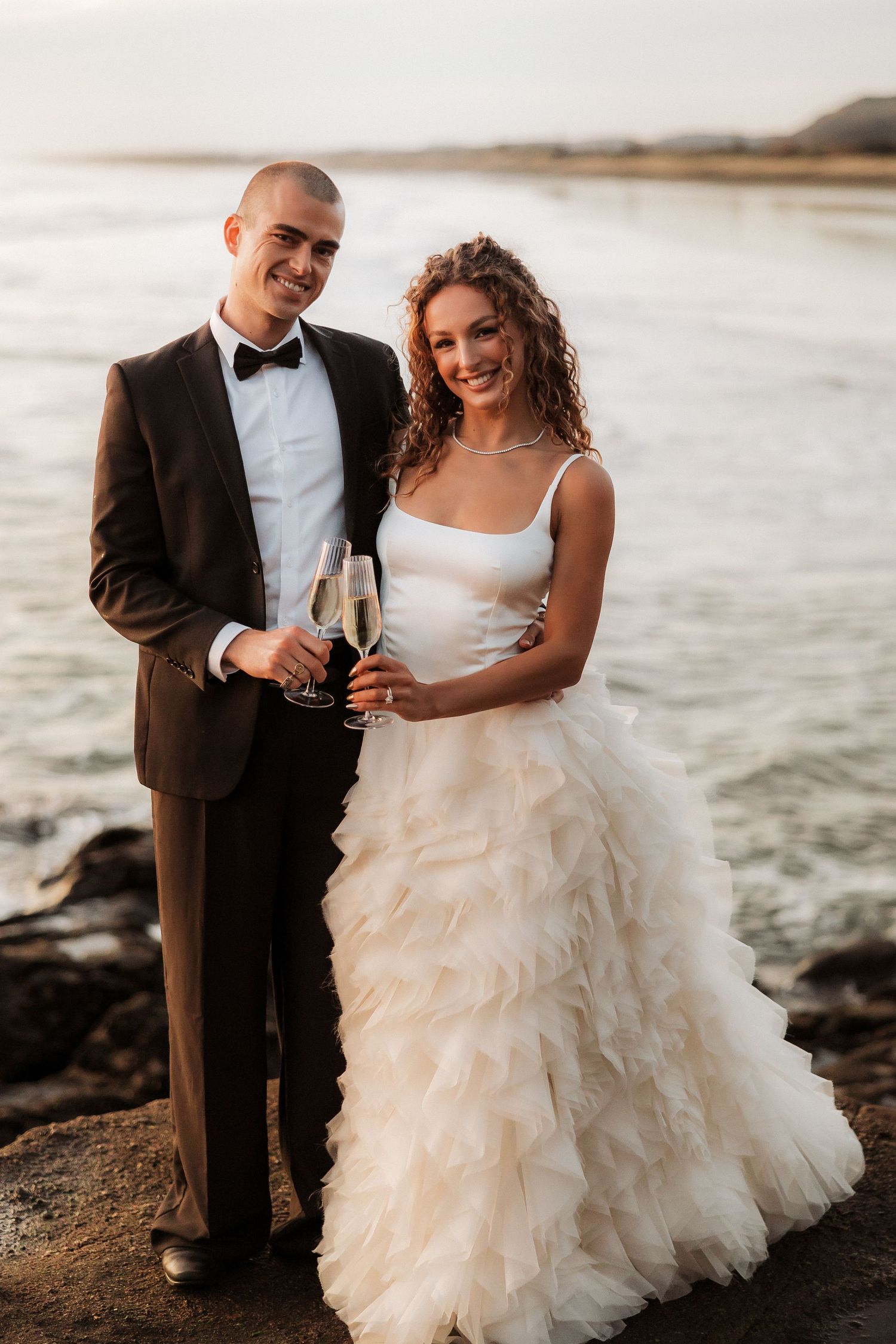 Wedding couple toasting with champagne glasses by the ocean at sunset.