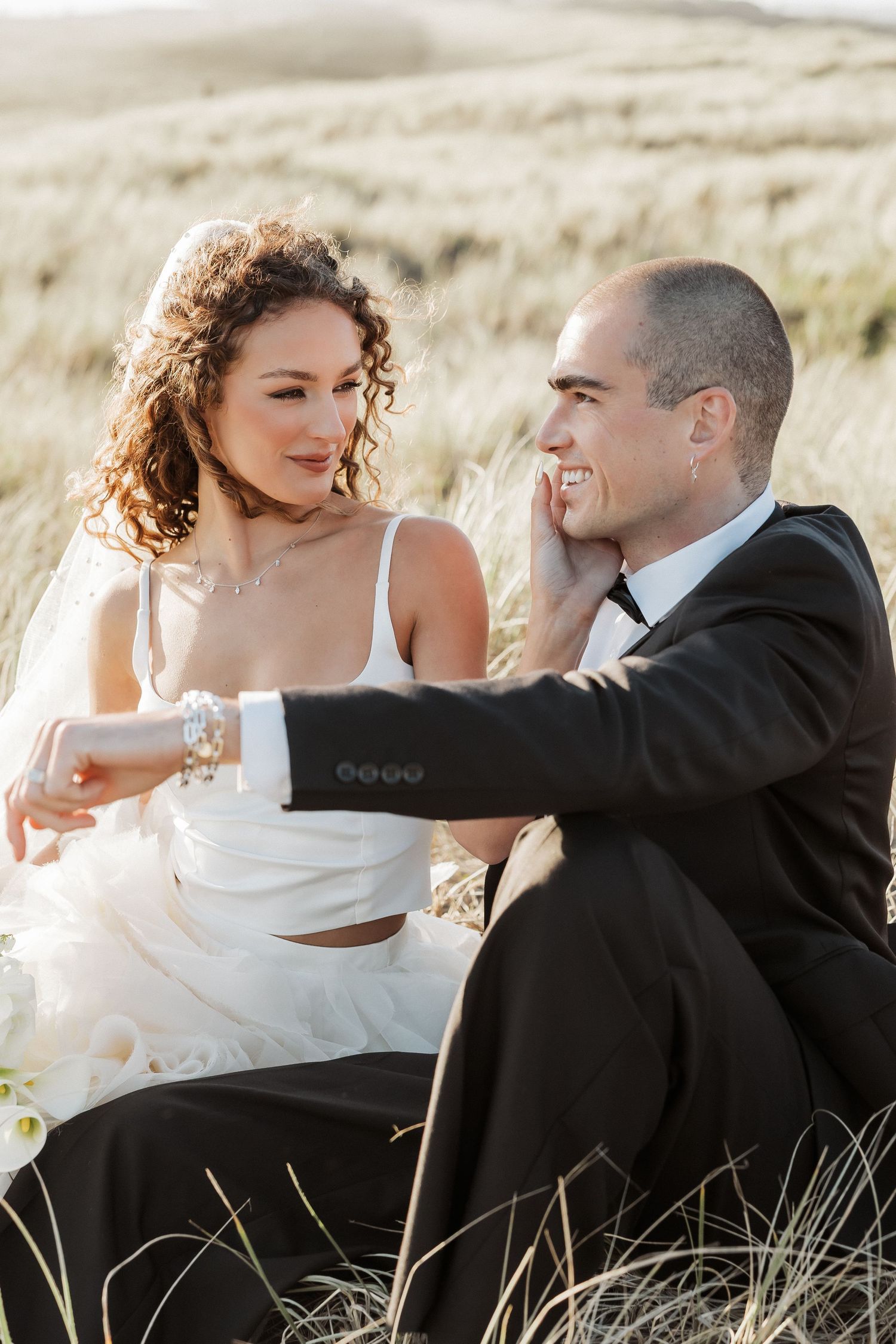 Romantic wedding portraits of newlyweds sitting together in golden grass at sunset.