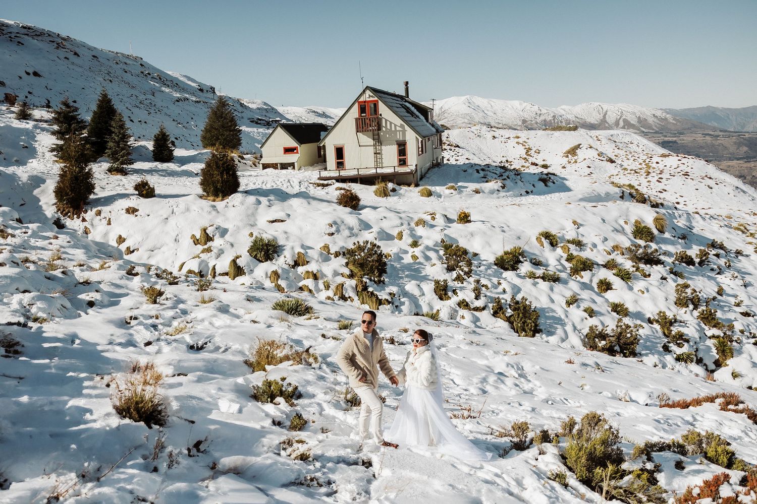 A snow-covered mountain cabin sits perched on a hillside with winter peaks in the distance.