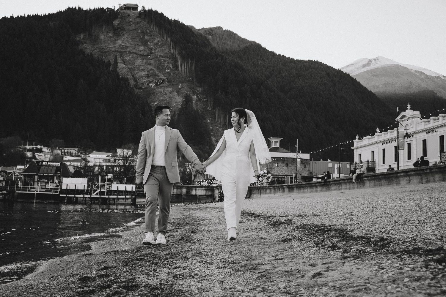 Black and white photo of couple walking along waterfront with mountains in background.