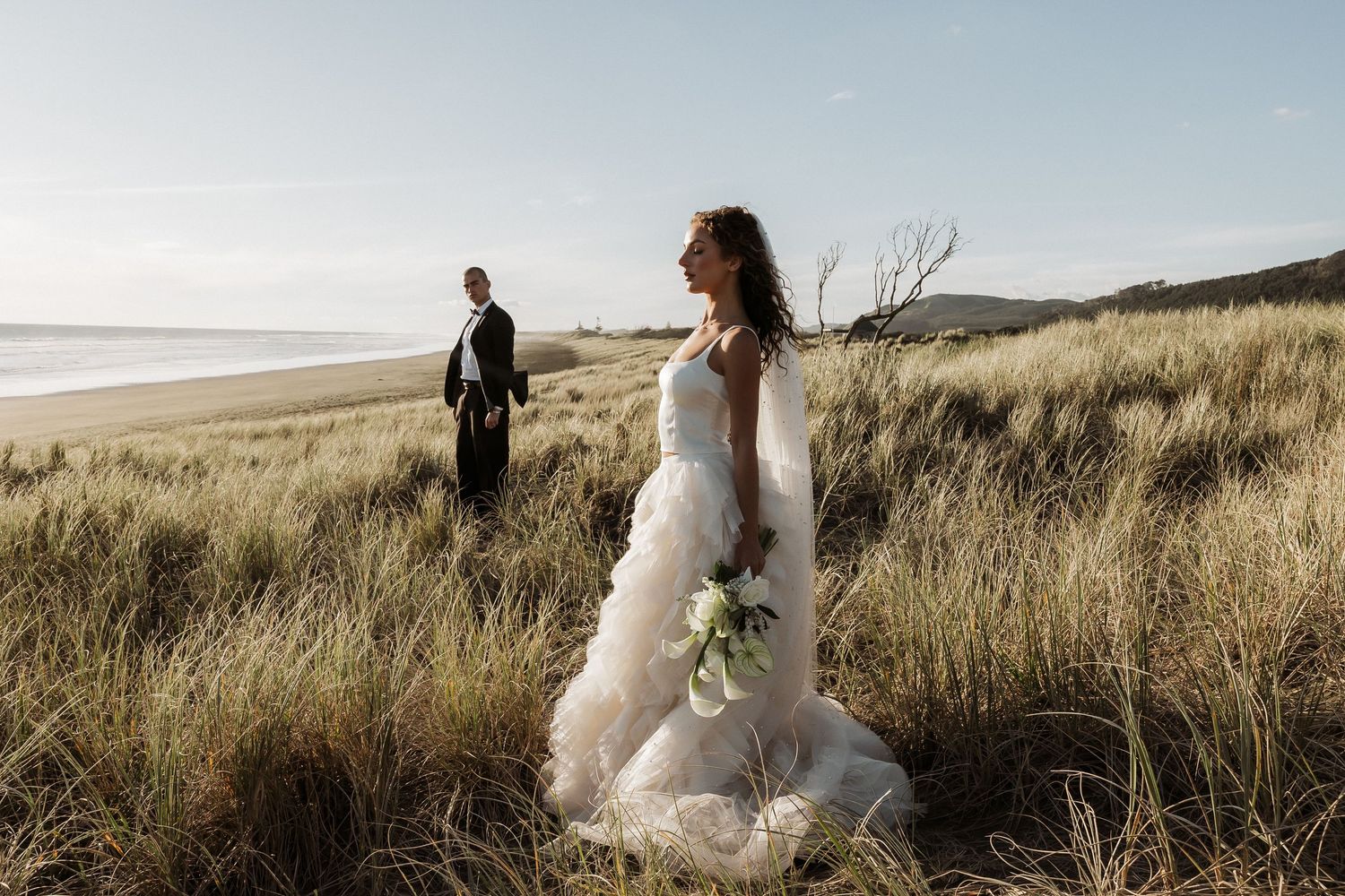 Bride in white ruffled gown stands in coastal grass dunes holding bouquet with ocean view.