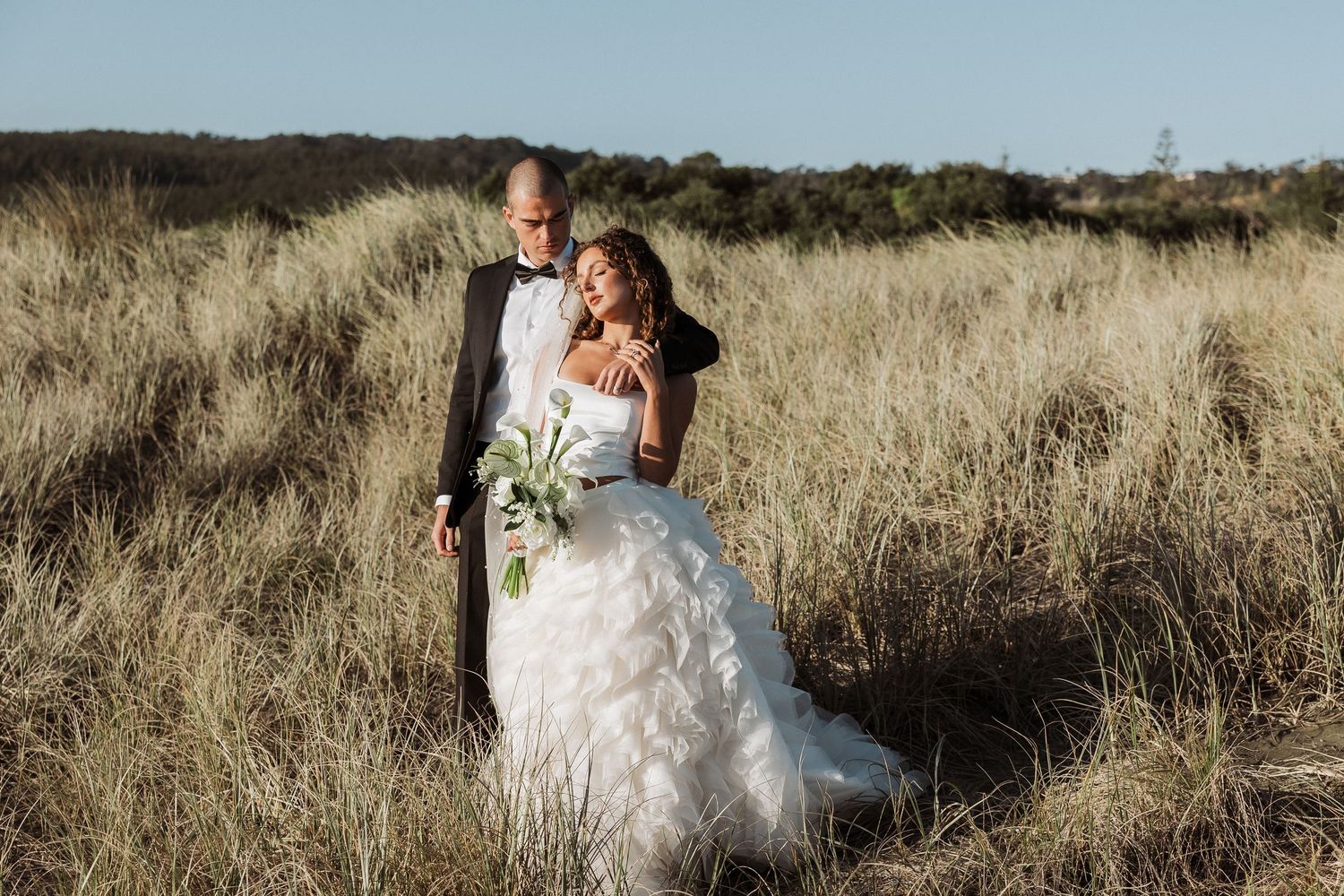 A romantic couple in wedding attire embraces while standing in a golden field at sunset.