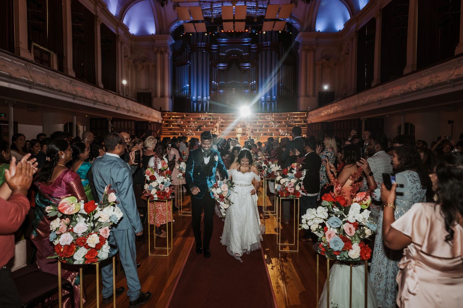 Wedding guests dance and celebrate on a dimly lit dance floor inside an elegant ballroom venue with arched ceilings.