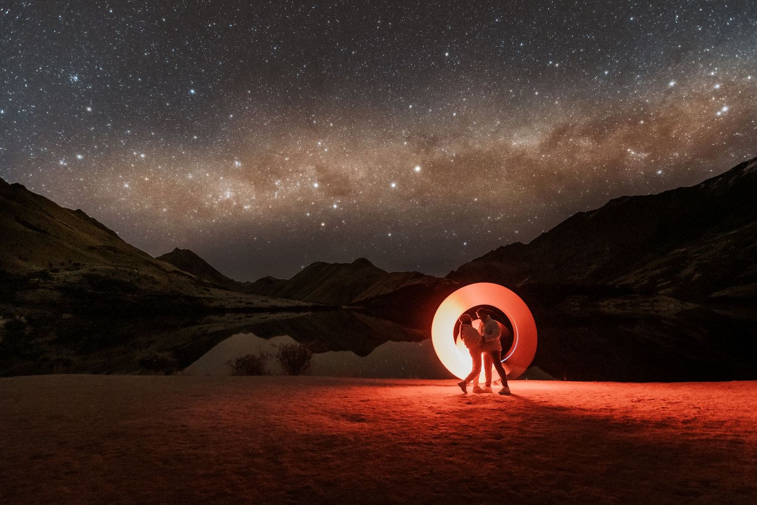 Light painting creates a glowing red circle against a starry night sky over a mountain lake landscape.
