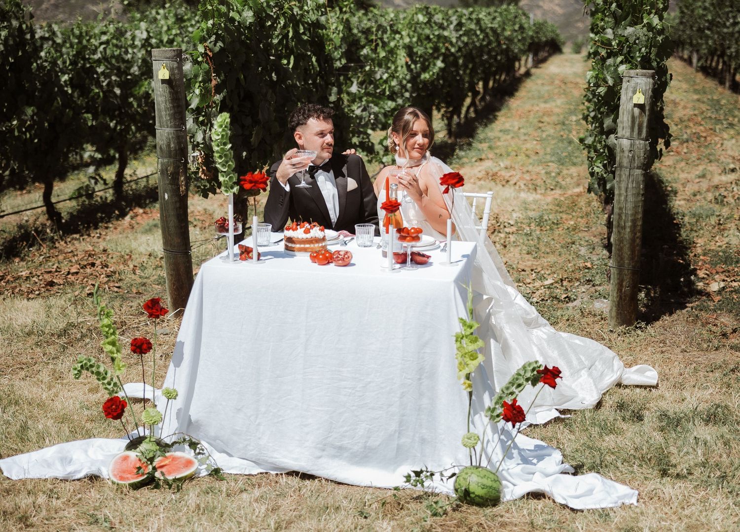 Romantic outdoor dining setup in a vineyard with white tablecloth and red floral decorations.