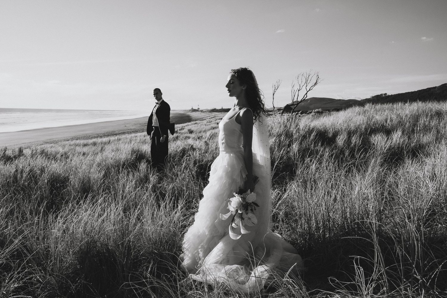 Couple sitting in tall golden grass at sunset on a hillside overlooking a scenic landscape.