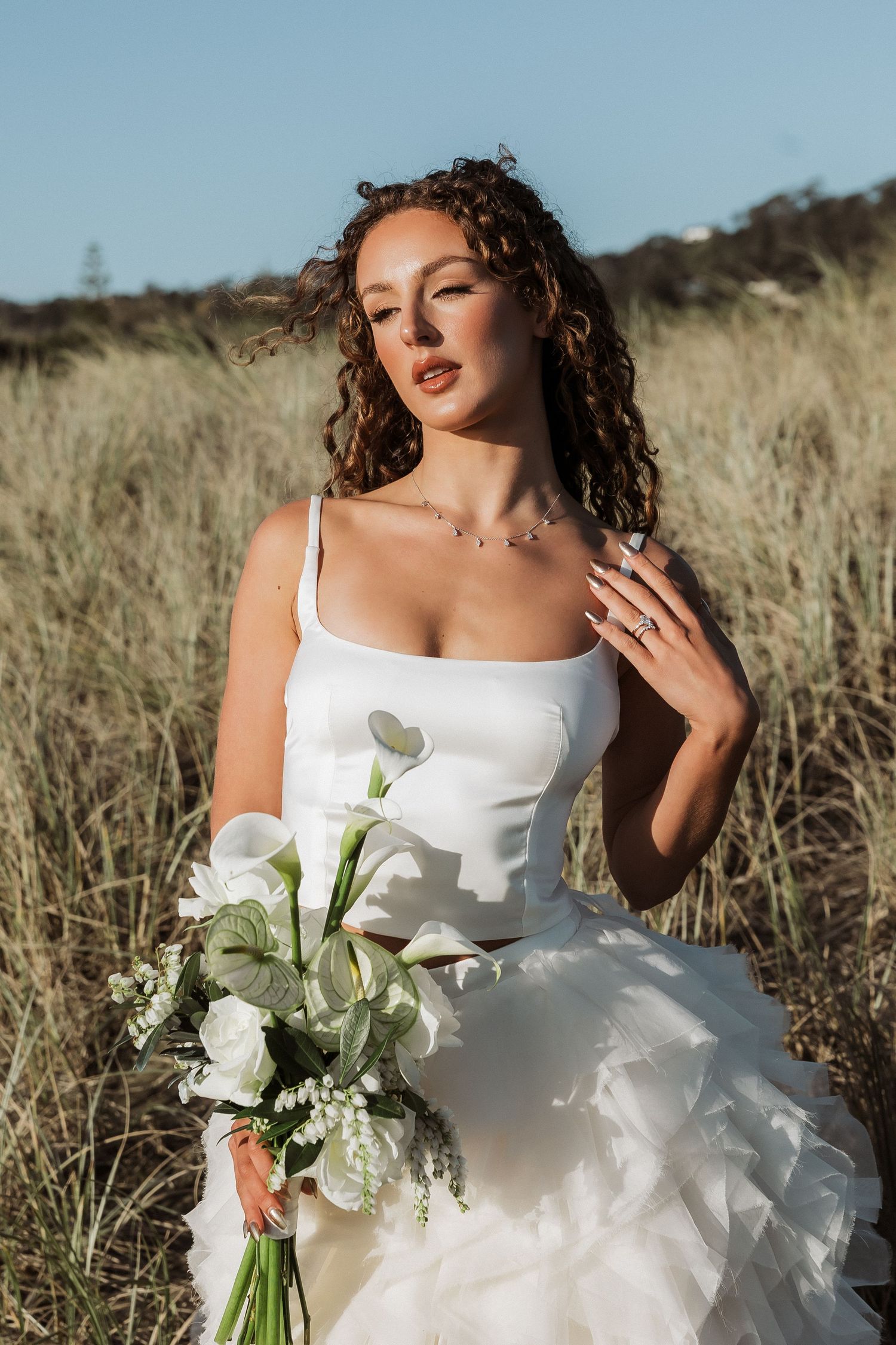 A bride in a white wedding dress holds a delicate bouquet of white flowers while posing in a sunlit field of tall grass.