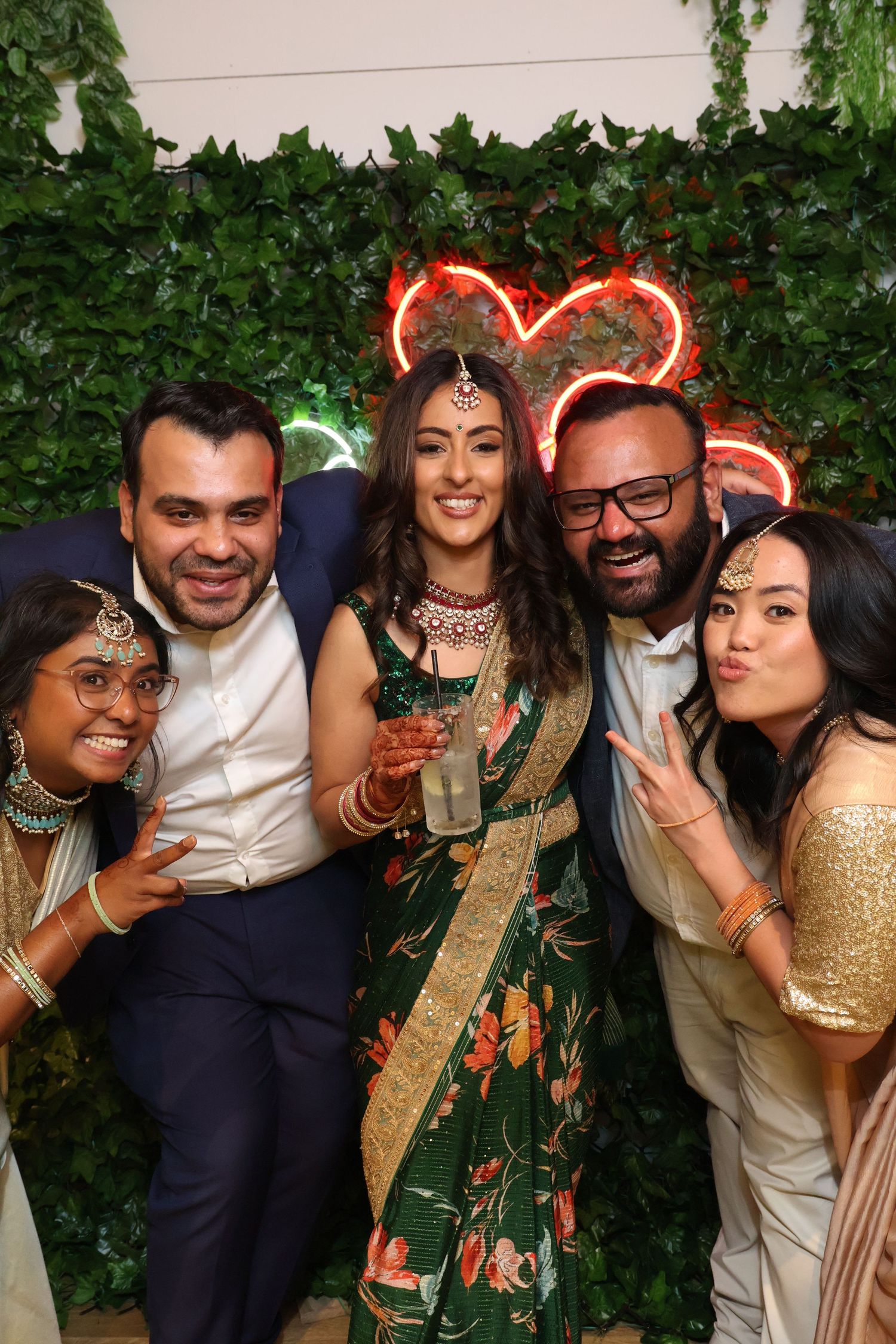 Friends celebrate at a party with a neon heart sign and ivy wall backdrop.