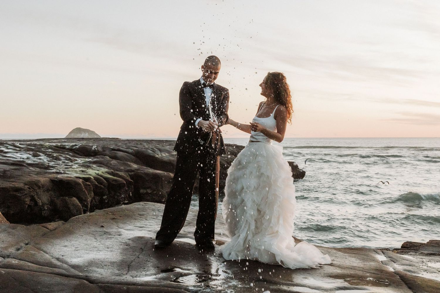 A couple stands on rocky coastal cliffs at sunset while ocean waves crash against the shore.