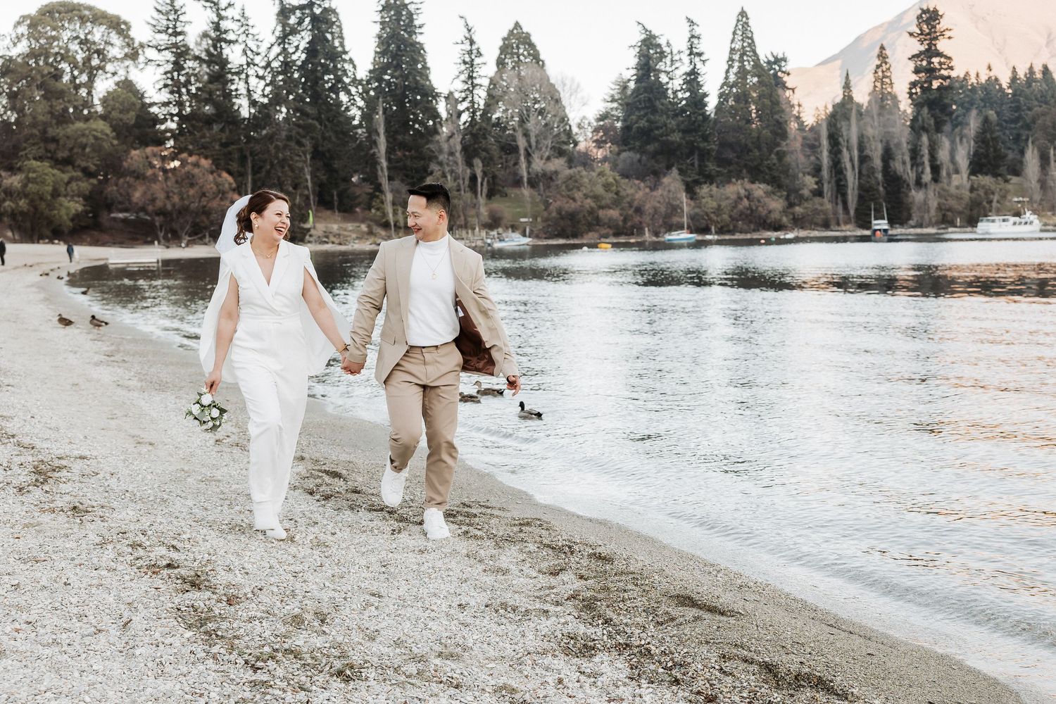 A couple walks hand in hand along a serene lakeshore with evergreen trees and mountains in the background.