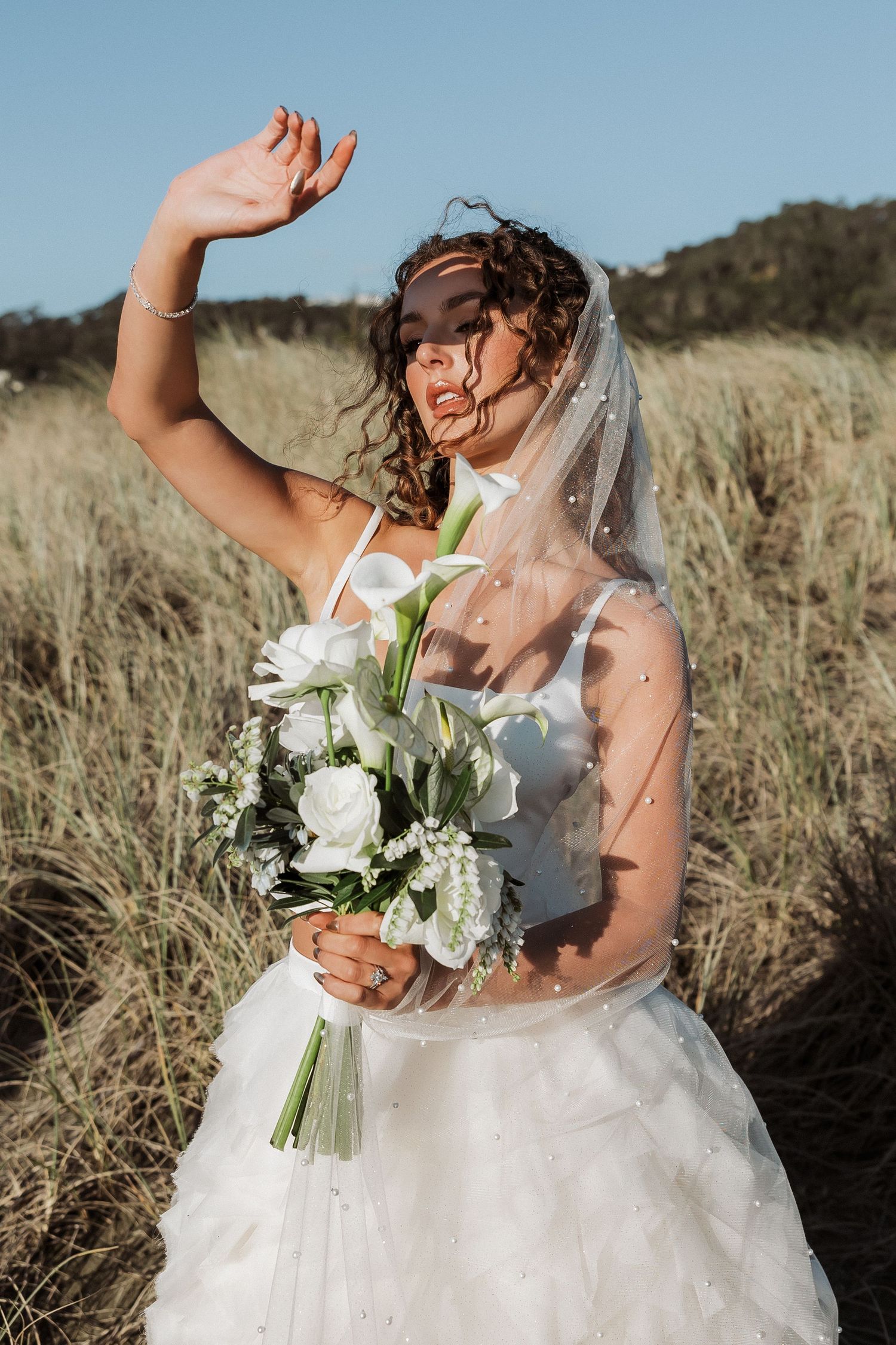 A joyful bride in a white gown dances and laughs while holding a bouquet of white flowers in a grassy field.