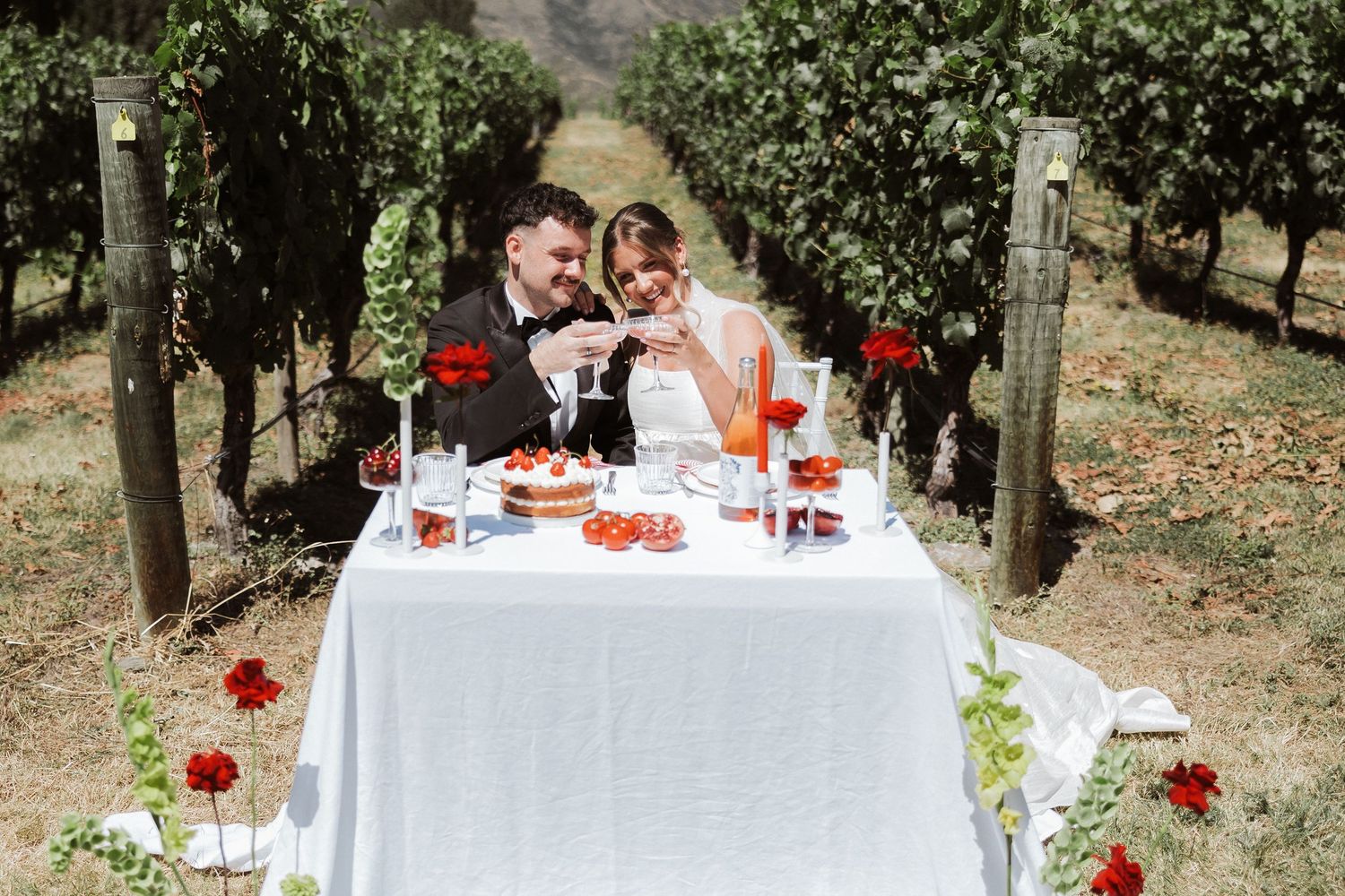 A romantic white table set for two with red florals and drinks in a vineyard setting.