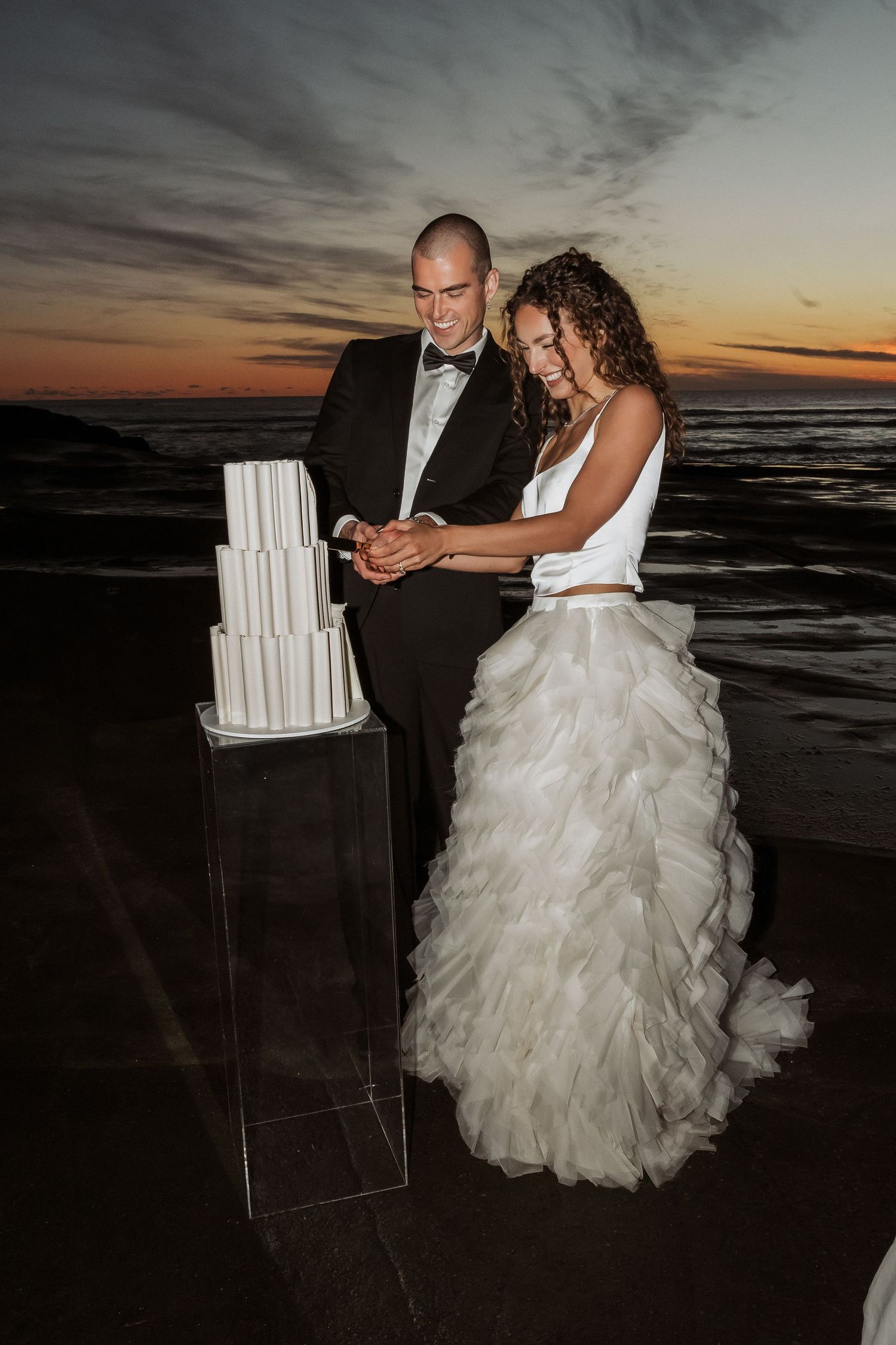 A wedding couple cuts their elegant white tiered cake on the beach at sunset with ocean waves in the background.