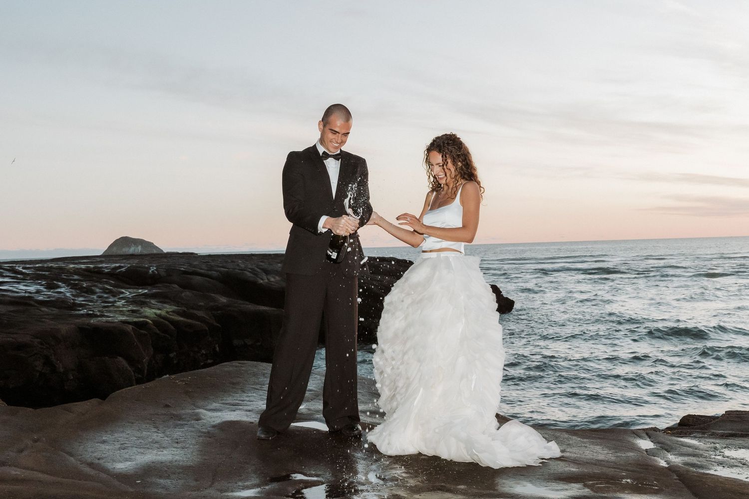 A couple in wedding attire stands on dramatic rocky cliffs overlooking the ocean at sunset.