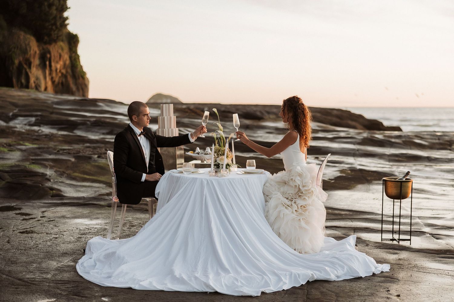 A romantic beach wedding scene with a couple sitting at an elegantly decorated table by the ocean at sunset.