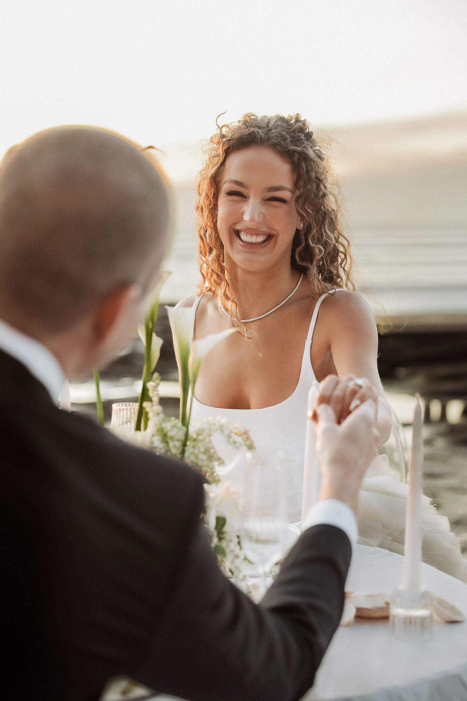 Wedding couple shares joyful laughter during outdoor reception by the ocean.