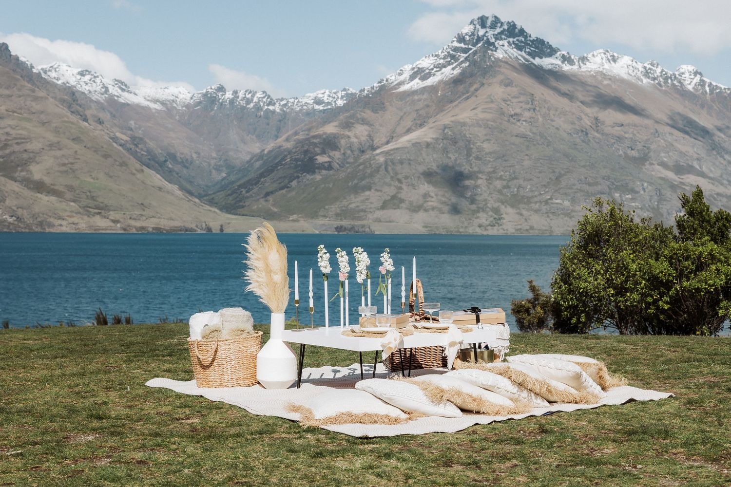 Elegant outdoor wedding setup with white decor and cushions against stunning mountain lake backdrop in New Zealand.