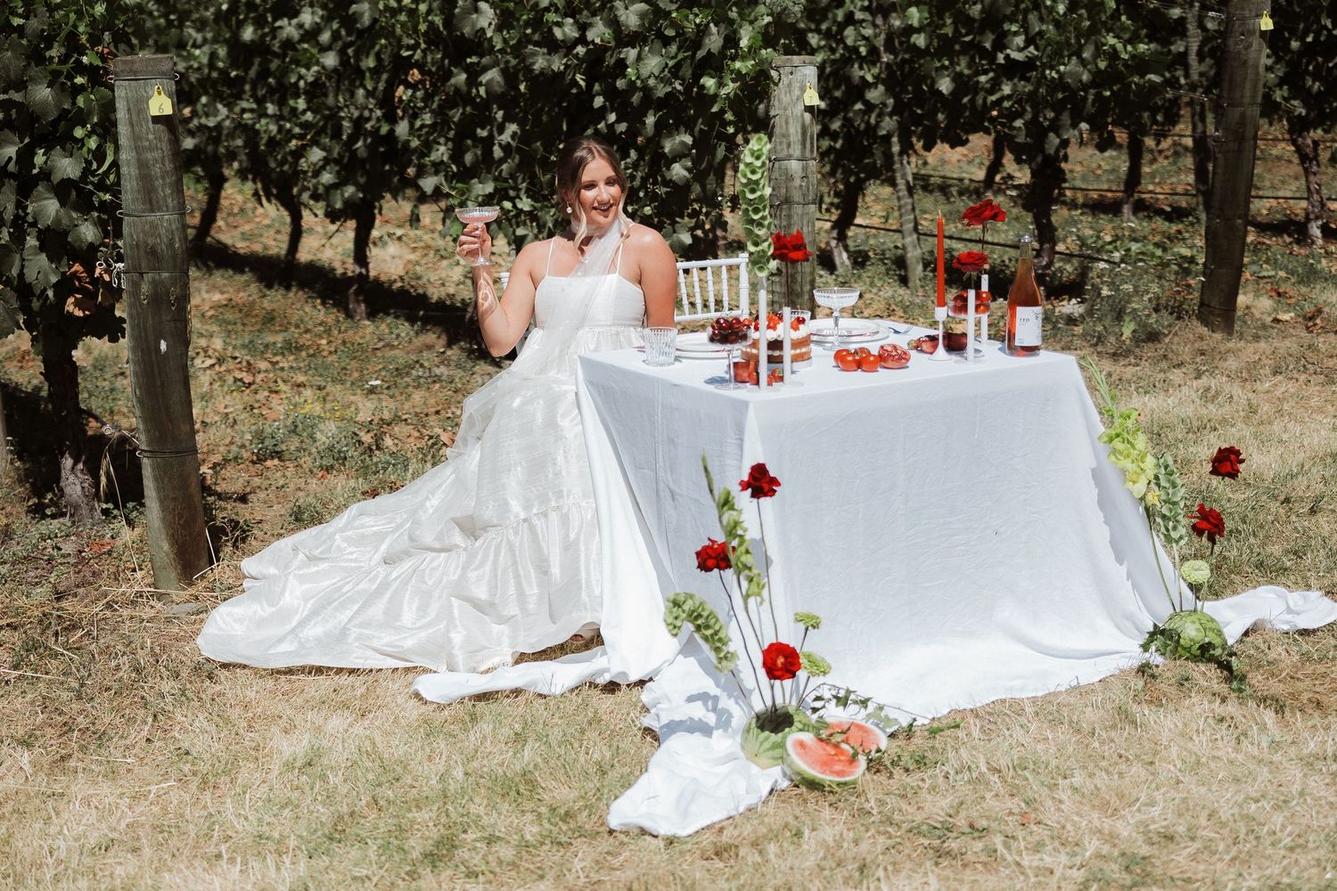 A bride in a flowing white gown poses gracefully on the ground surrounded by red roses and trees in a garden setting.