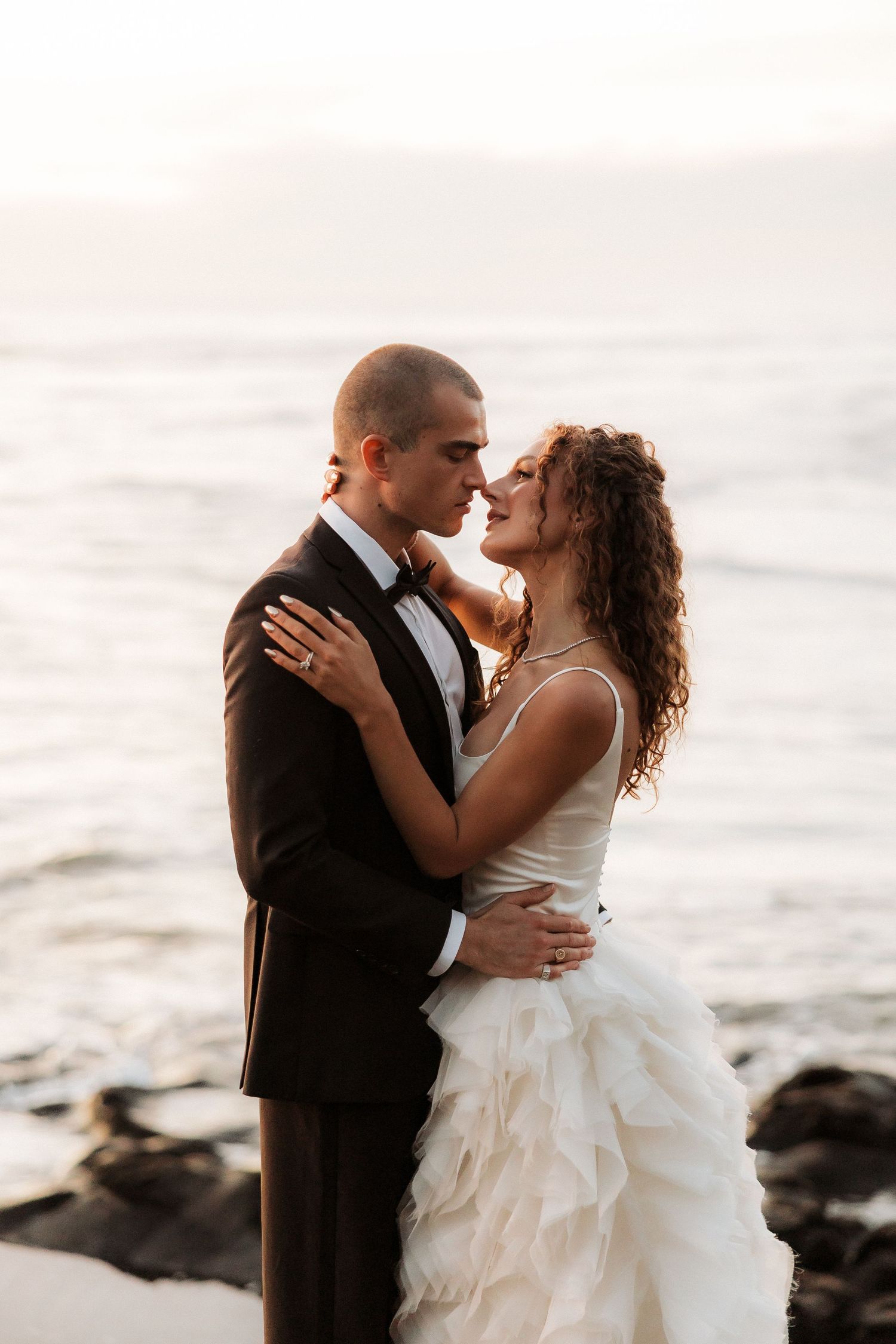 A romantic wedding couple embraces on a rocky beach at sunset with waves crashing behind them.