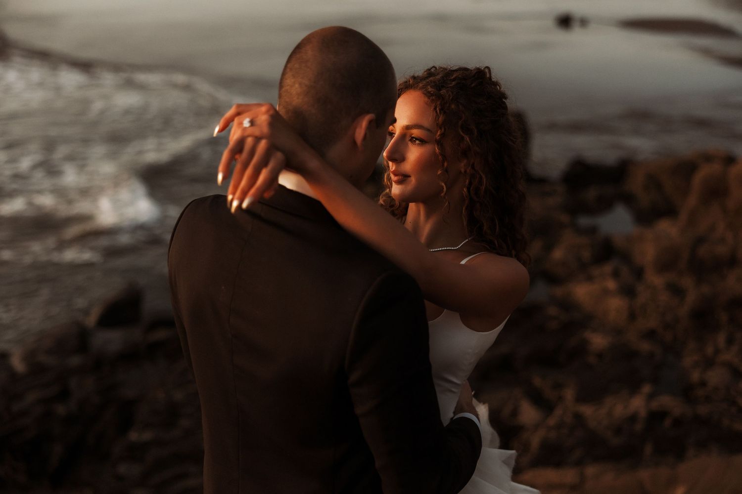 A romantic couple embraces on a rocky beach during golden hour as waves roll in behind them.