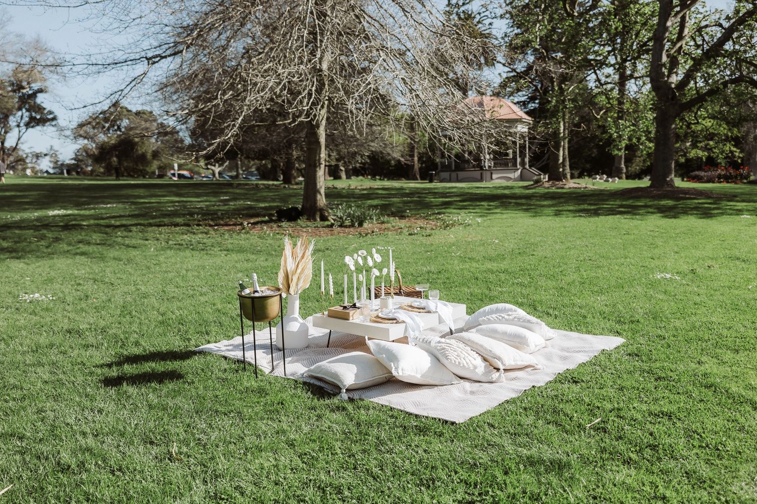A romantic picnic setup with white blankets and champagne on a lush green lawn near a historic house.