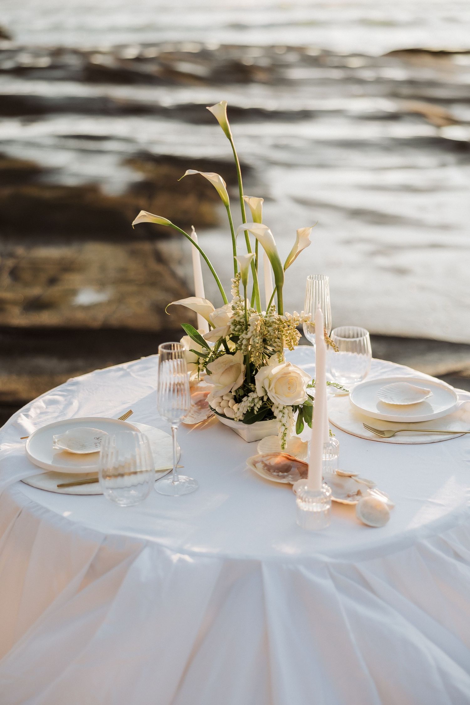 Elegant wedding table settings with white linens, calla lilies, and delicate glassware arranged along a beachside location.