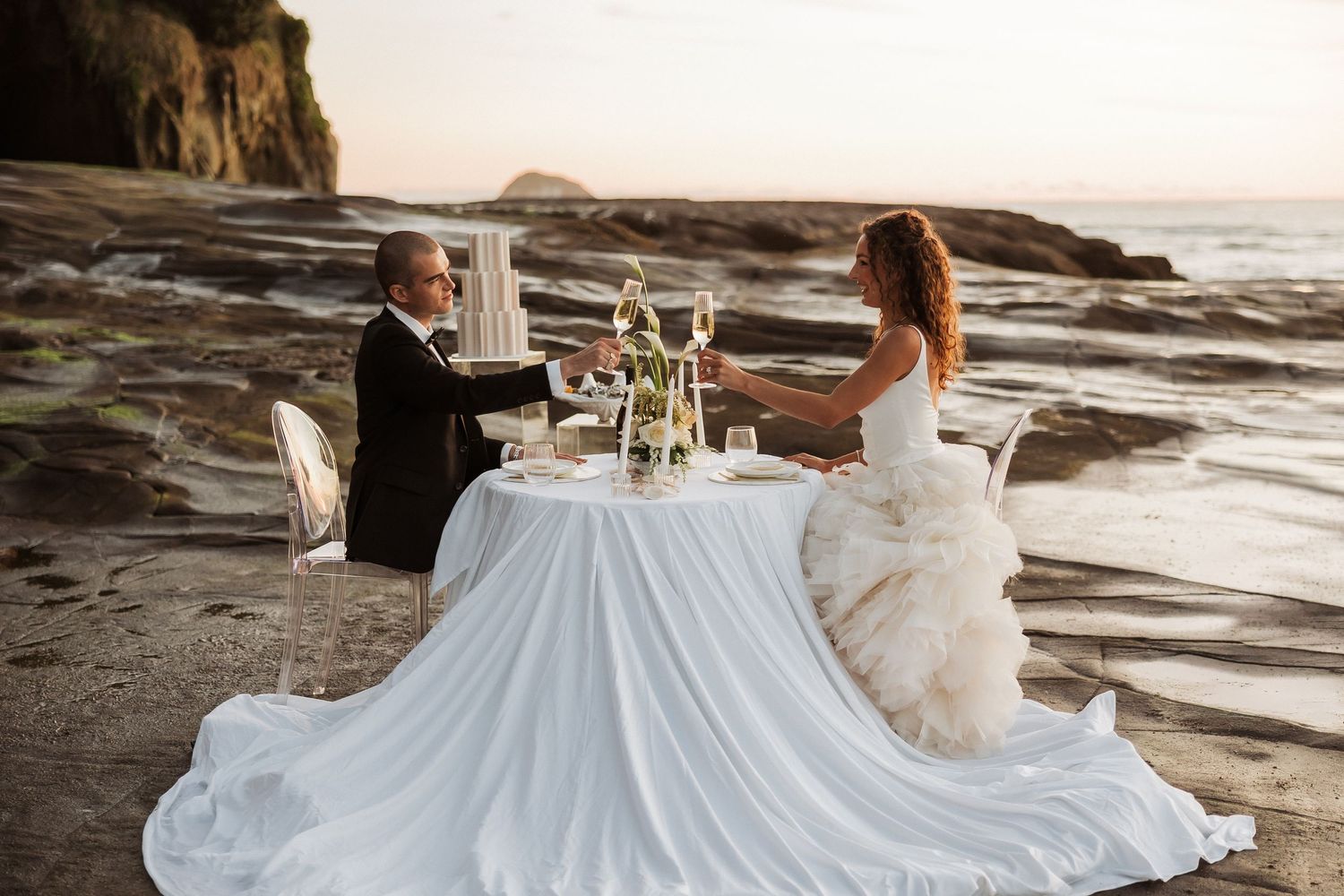 A romantic wedding dinner setup on rocky coastal cliffs at sunset with white tablecloth and elegant place settings.