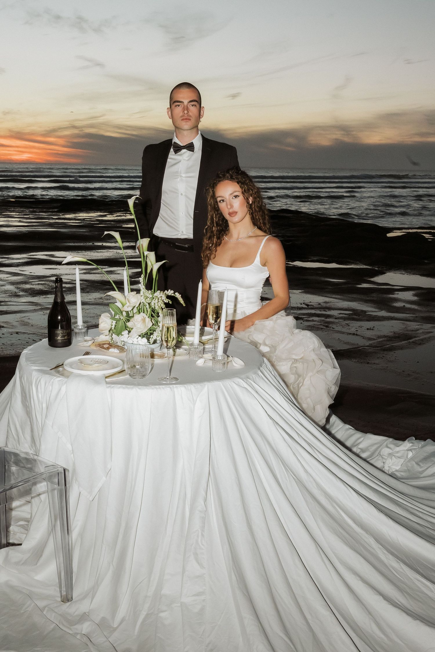 A wedding couple enjoys a romantic sunset dinner at a beachside table decorated with white linens and floral centerpieces.