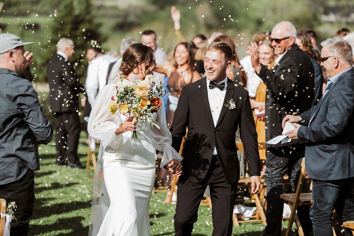 Wedding guests throw confetti at a joyful outdoor celebration as newlyweds walk past on a sunny day.