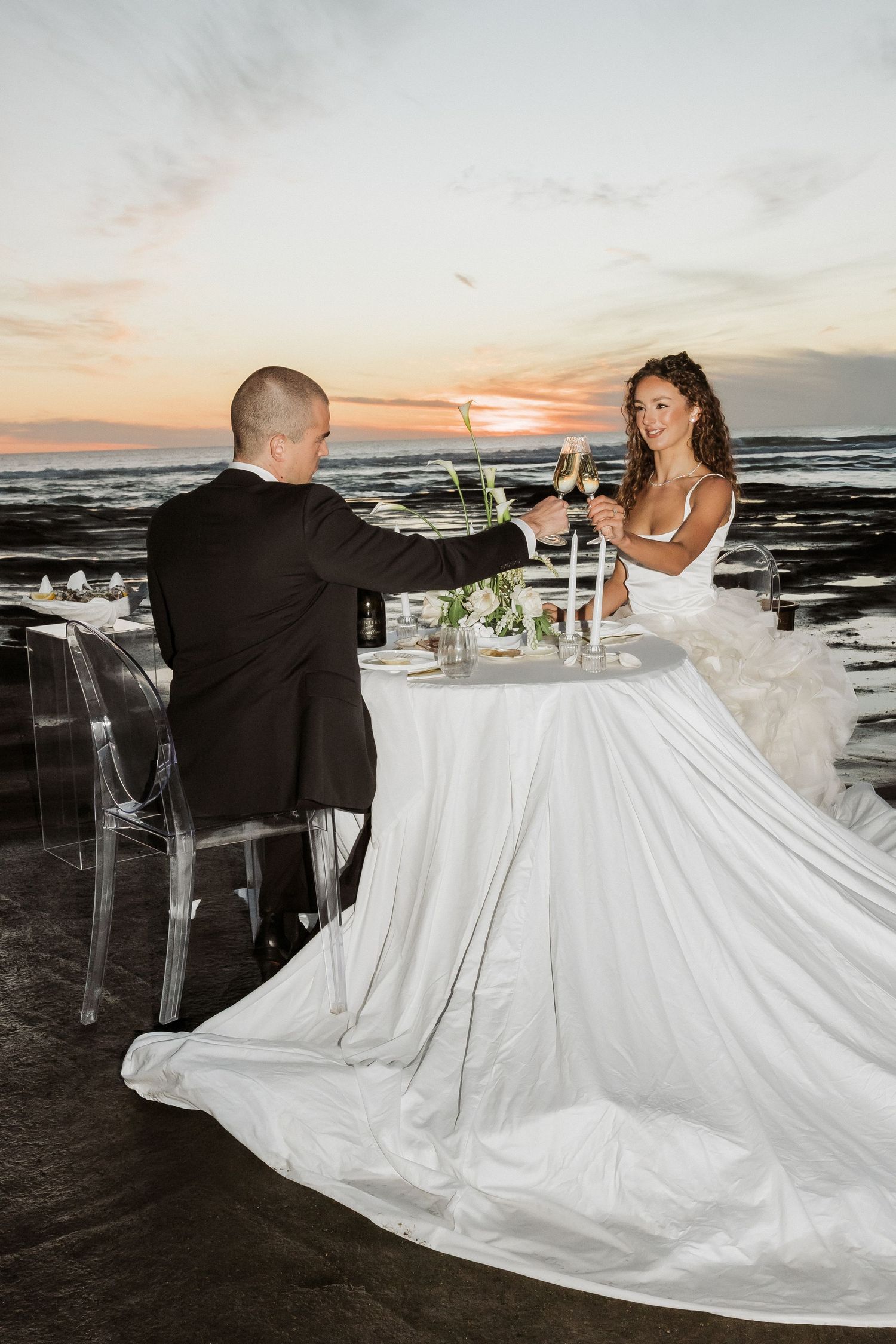 A couple sits at an elegant outdoor reception table overlooking the ocean at sunset, with flowing white wedding dress details.