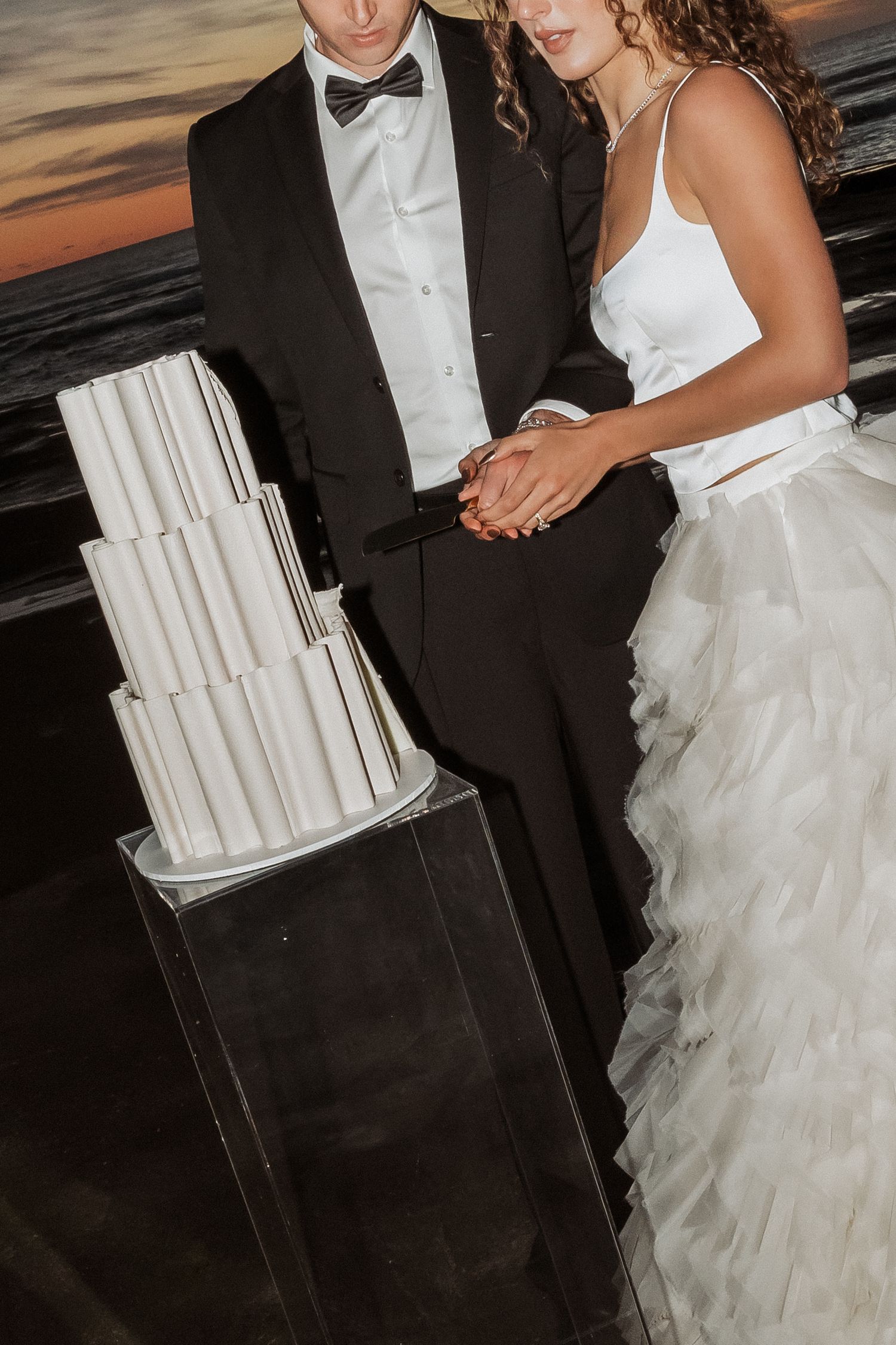 A bride and groom stand beside their modern white tiered wedding cake at sunset.