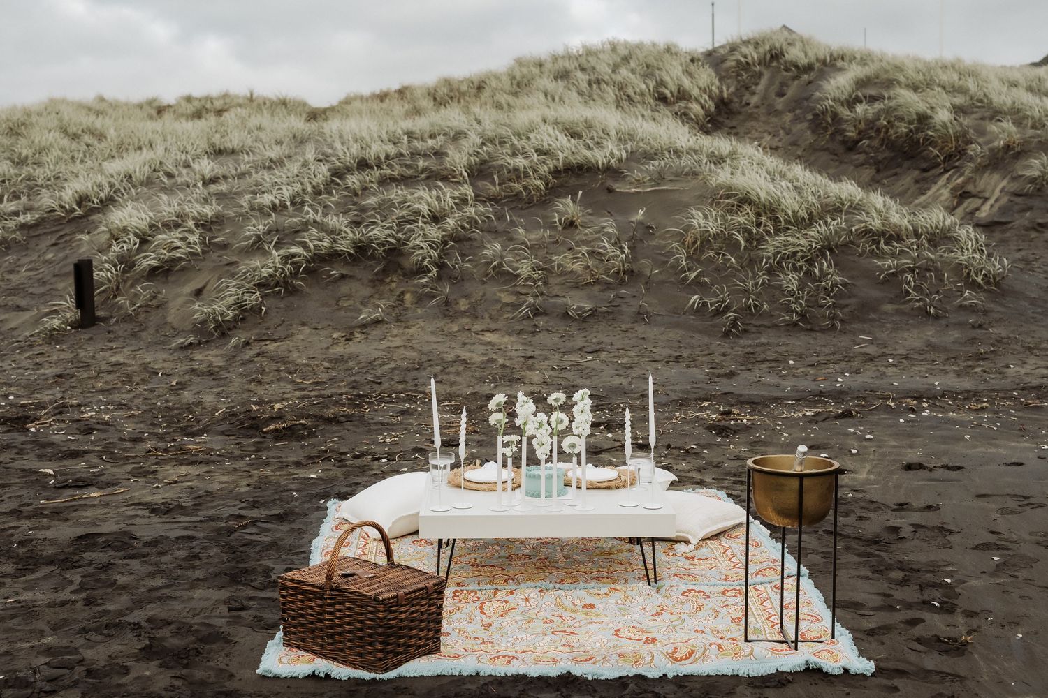 A series of rustic picnic tables with white tablecloths set up on a sandy beach against a cloudy sky.
