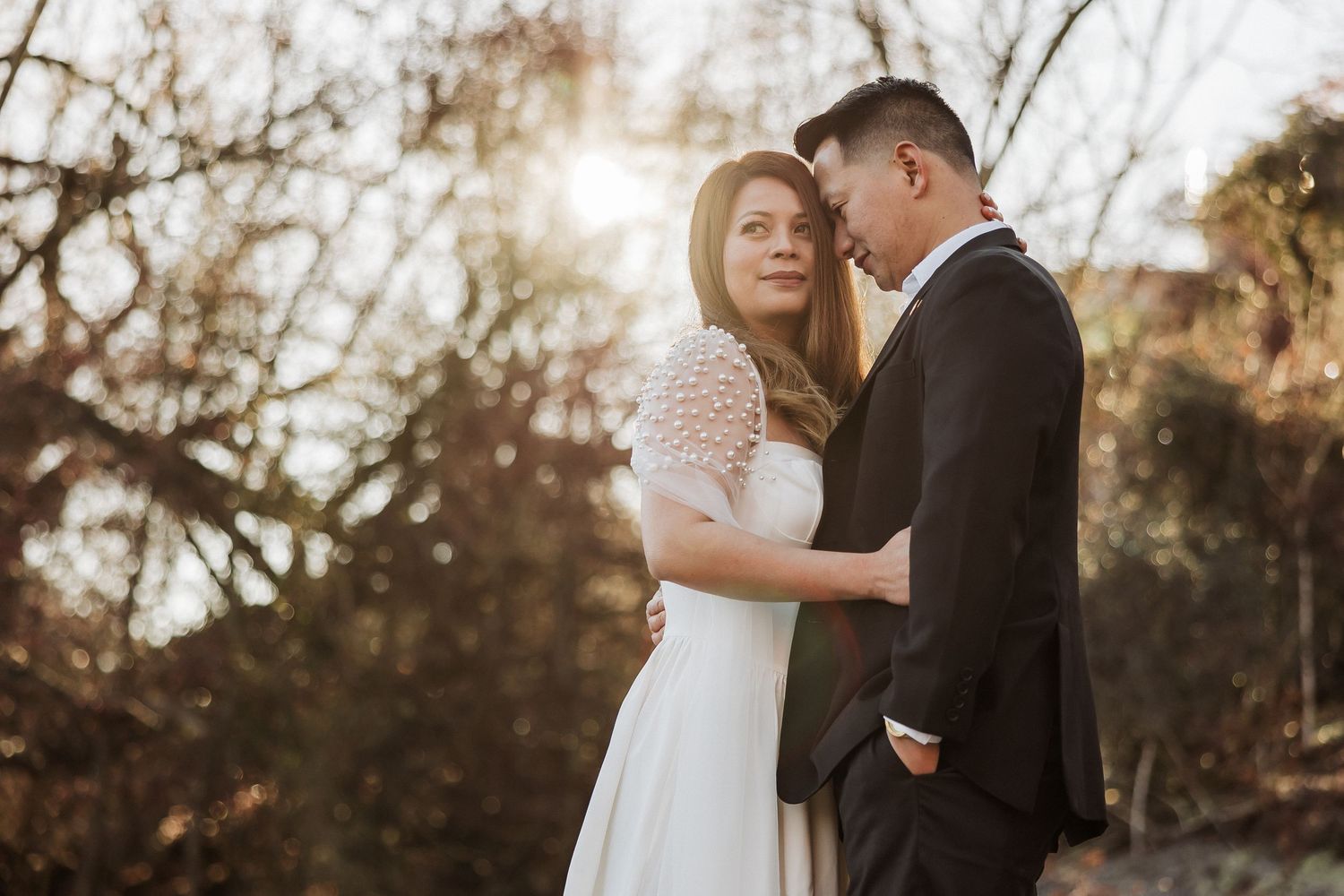 A couple in wedding attire shares an intimate moment outdoors during golden hour with trees in the background.